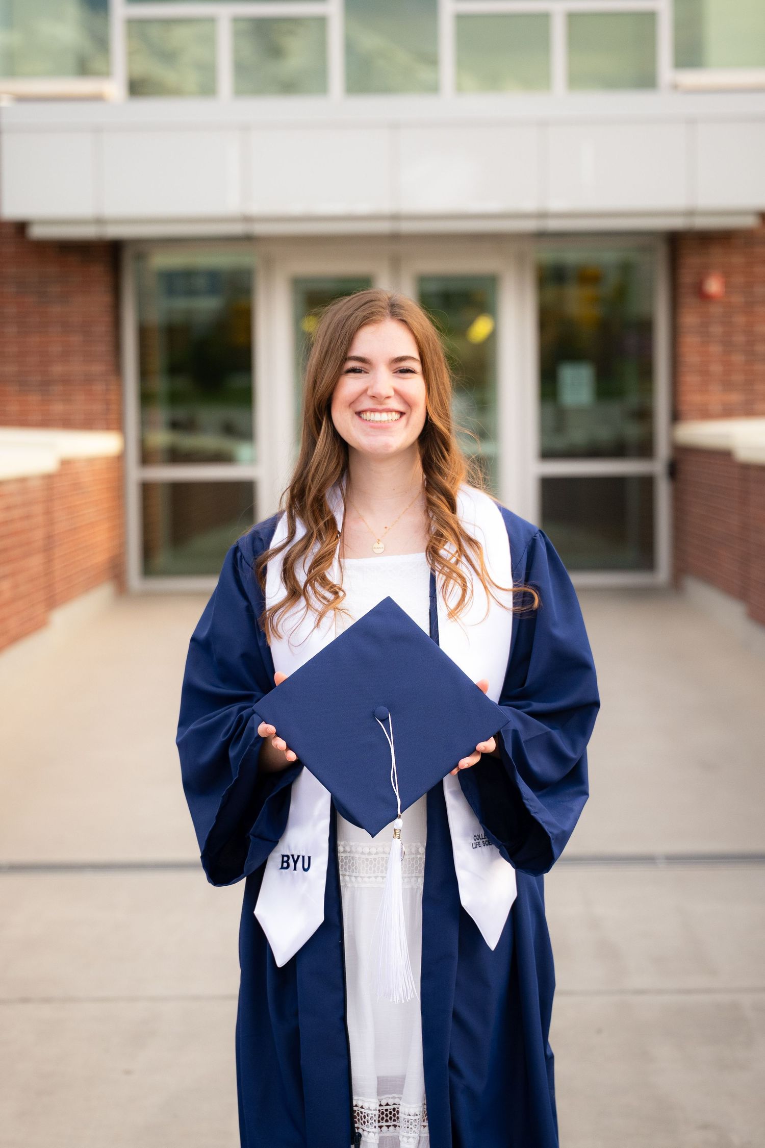 A young woman with long curly brown hair smiles while wearing a white dress and blue graduation gown in front of a building.