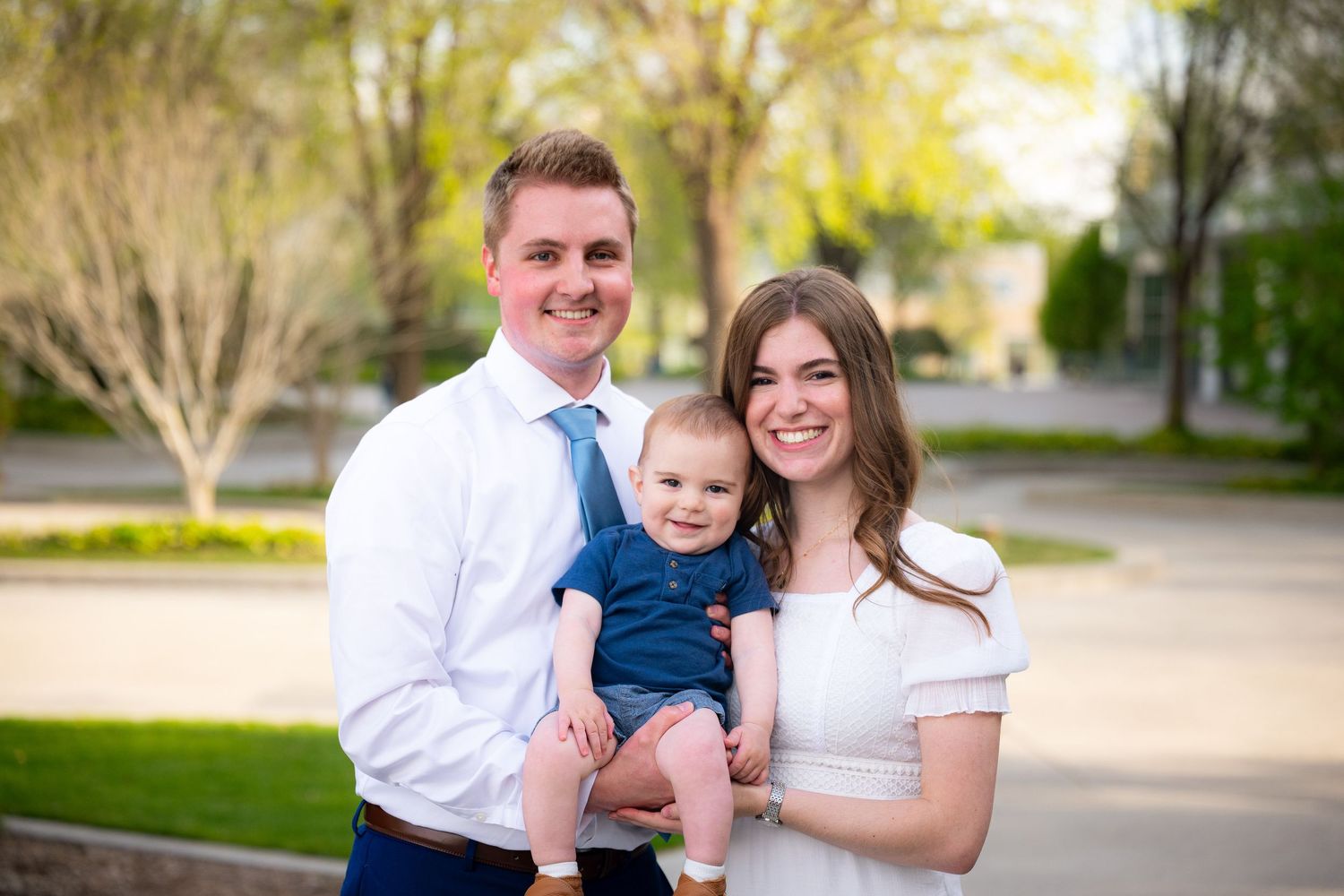 A family of three, including a baby, pose for a portrait outdoors.