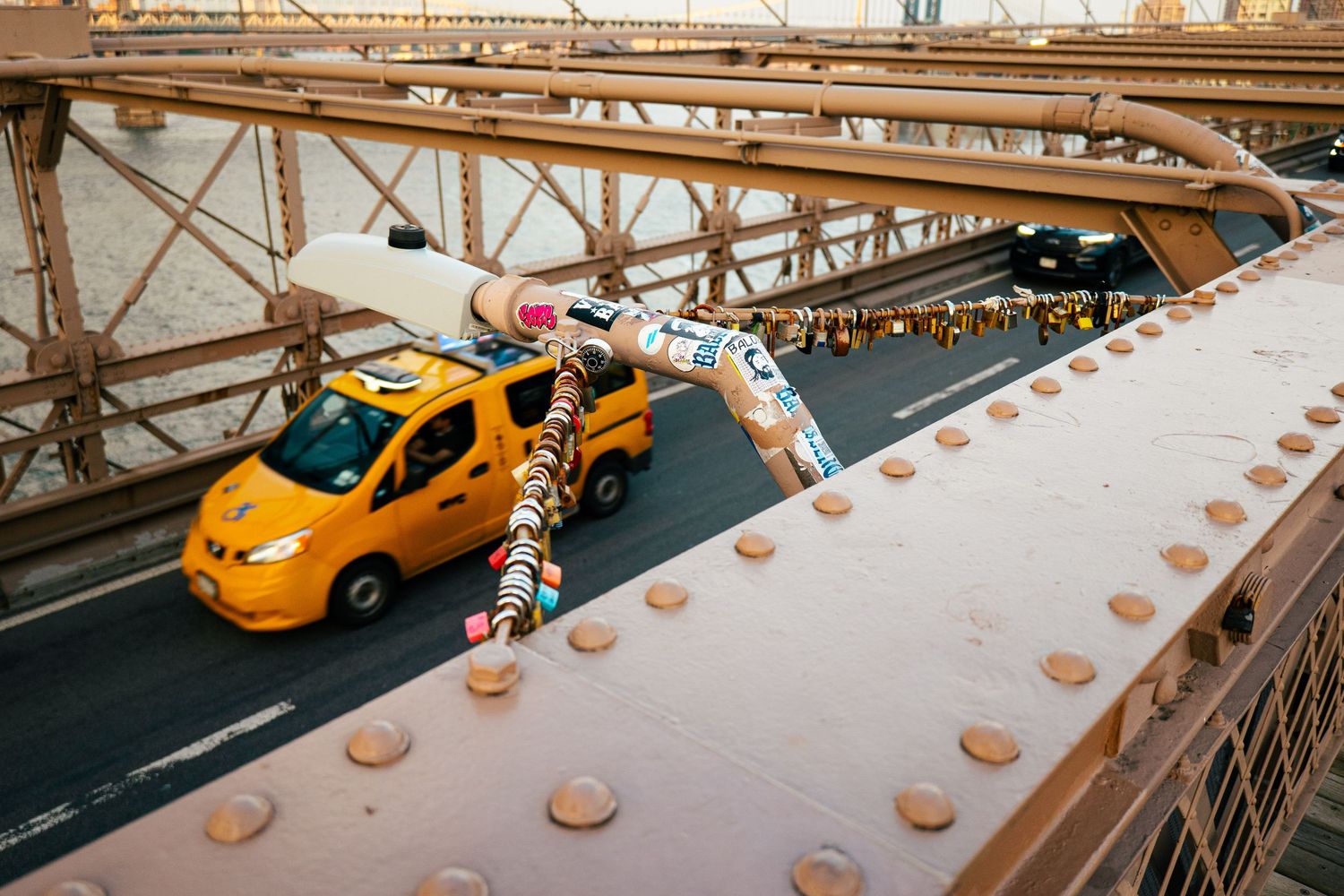 A yellow taxi cab driving across a historic suspension bridge in an urban setting.