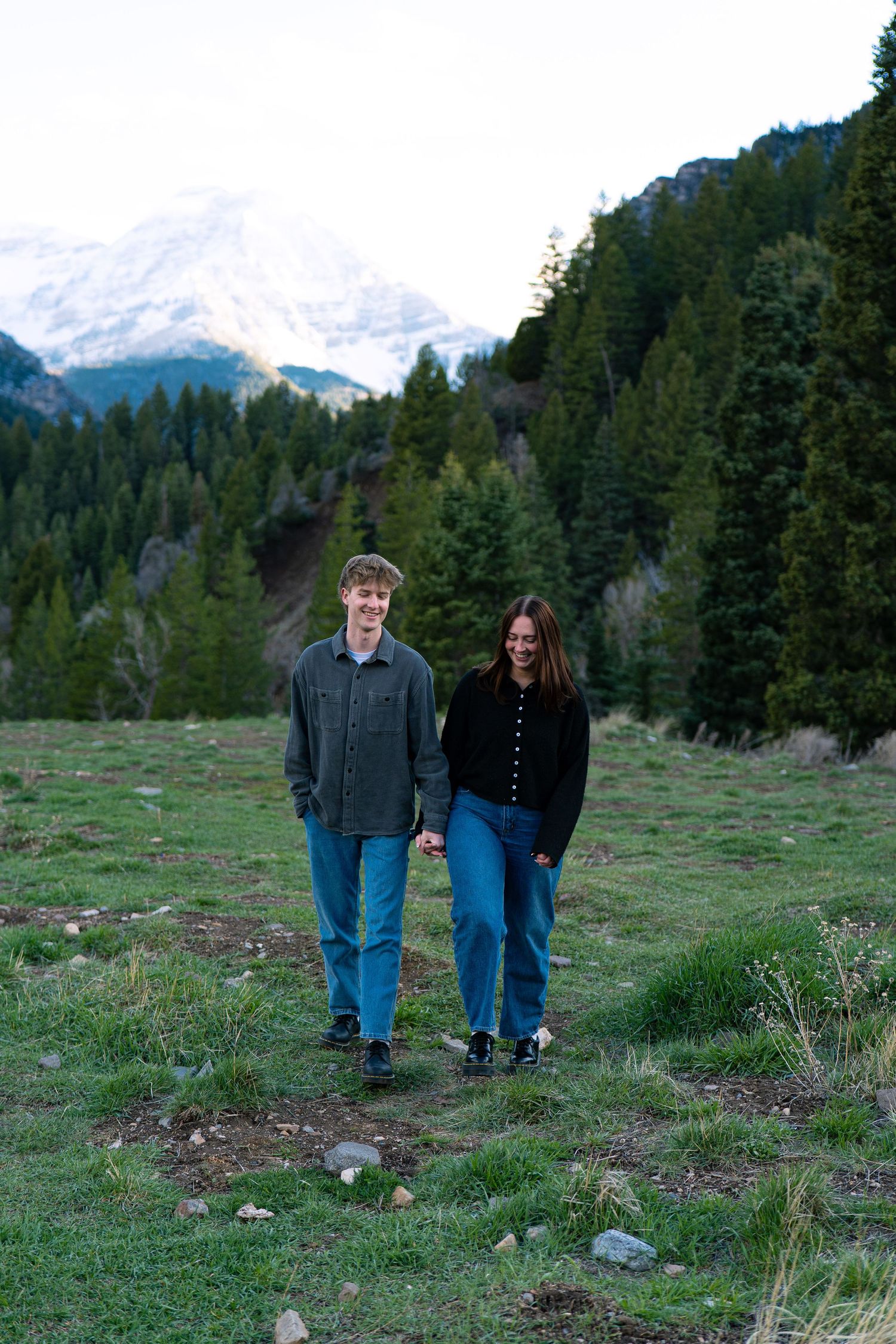 Couple holding hands walking on a grassy mountain trail with snow-capped peaks and pine trees.