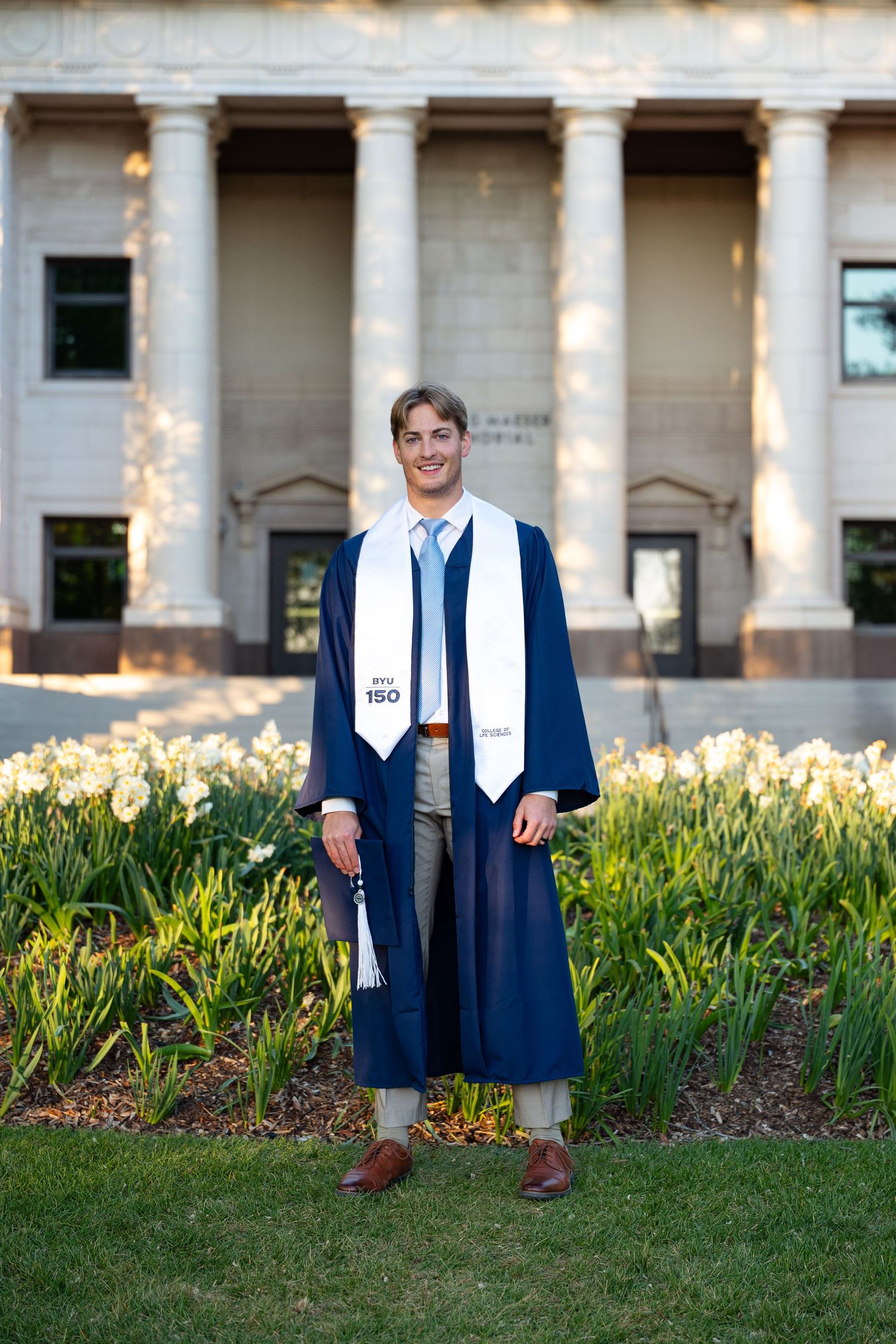Young man in navy graduation gown and white stole smiles outdoors near spring flowers and classical building columns.