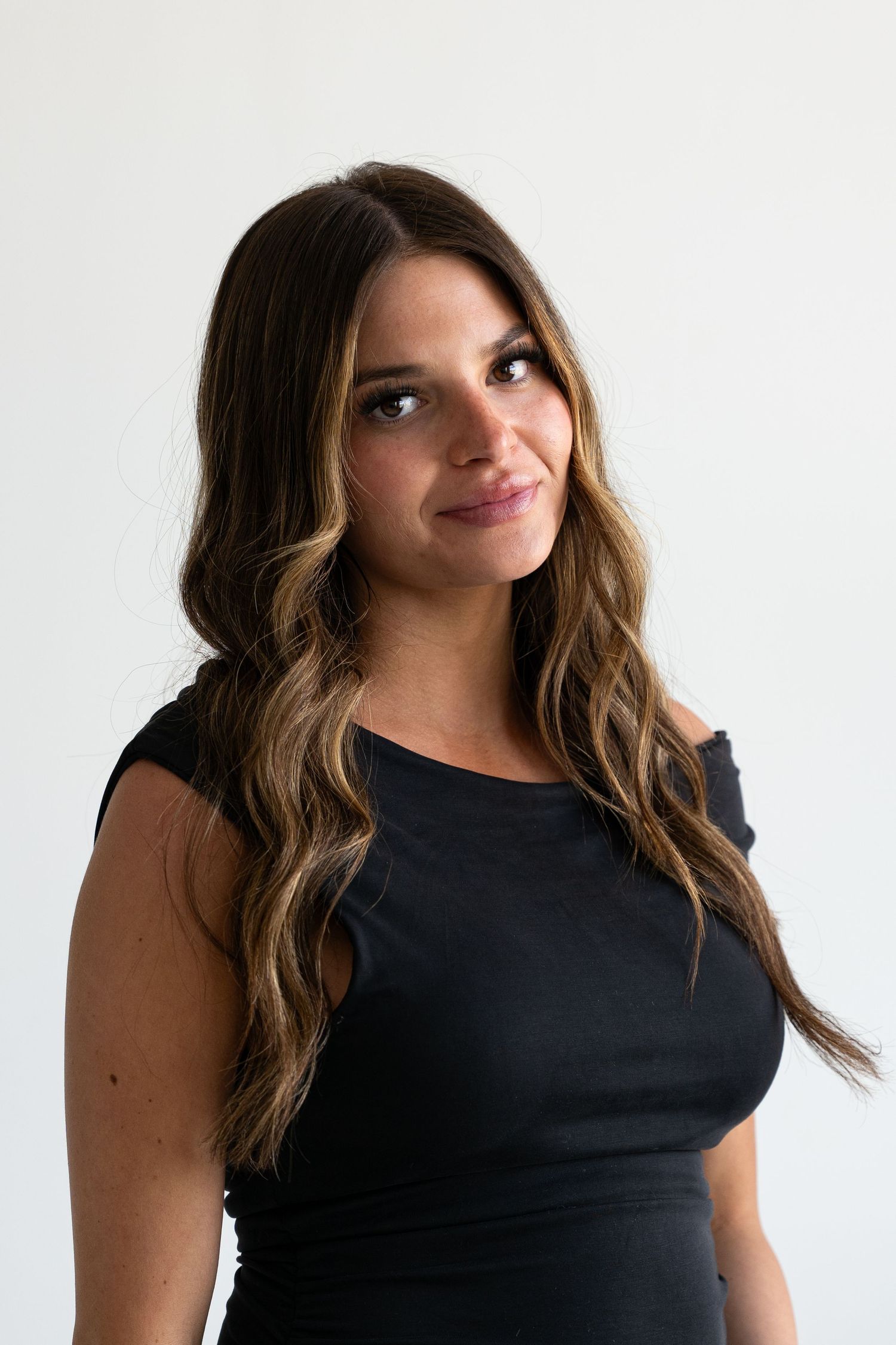 A young woman with long wavy brunette hair smiles softly, wearing a black sleeveless top against a white background.