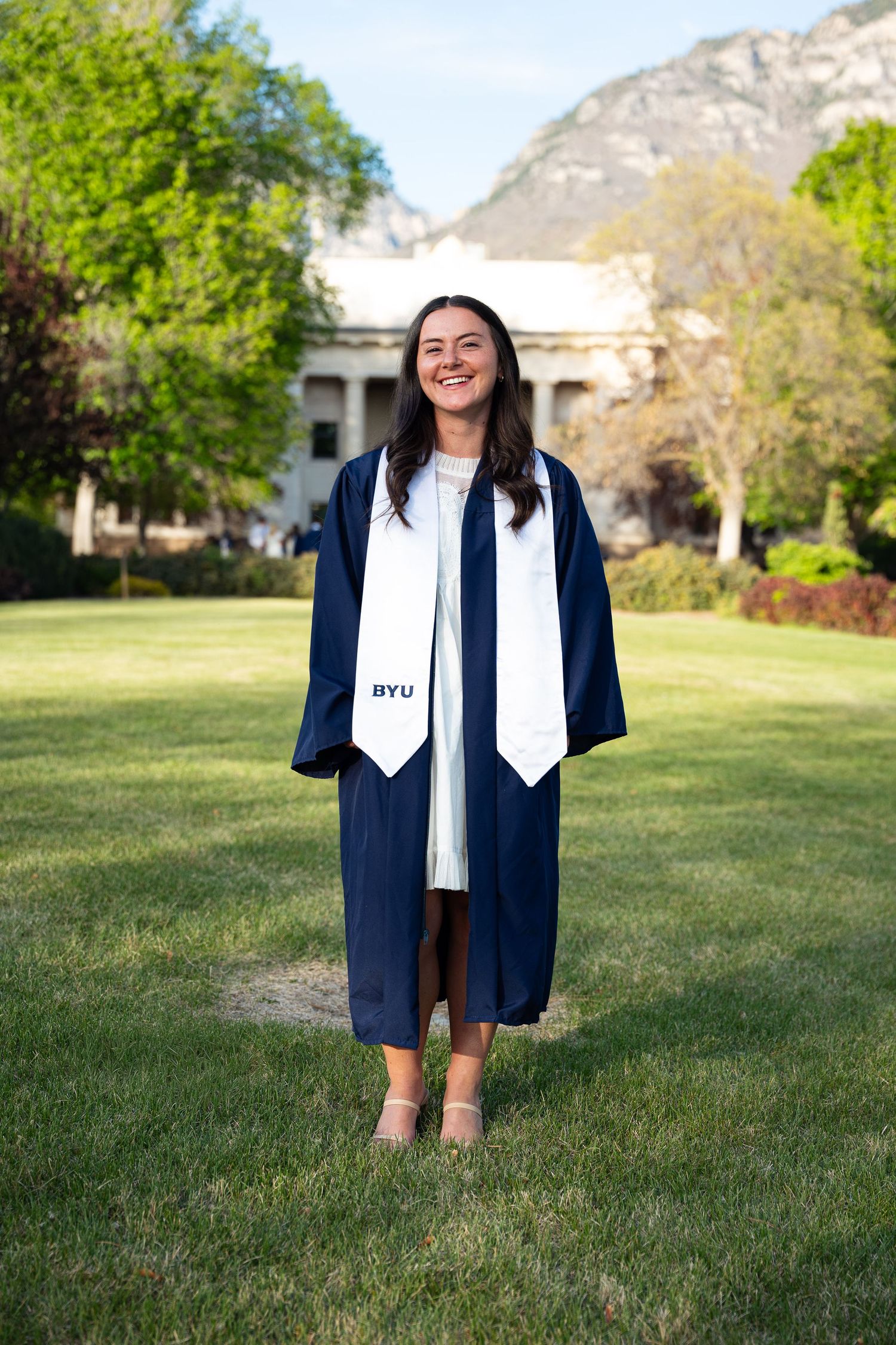 BYU graduate smiling in navy cap and gown on green campus lawn with mountains in background.