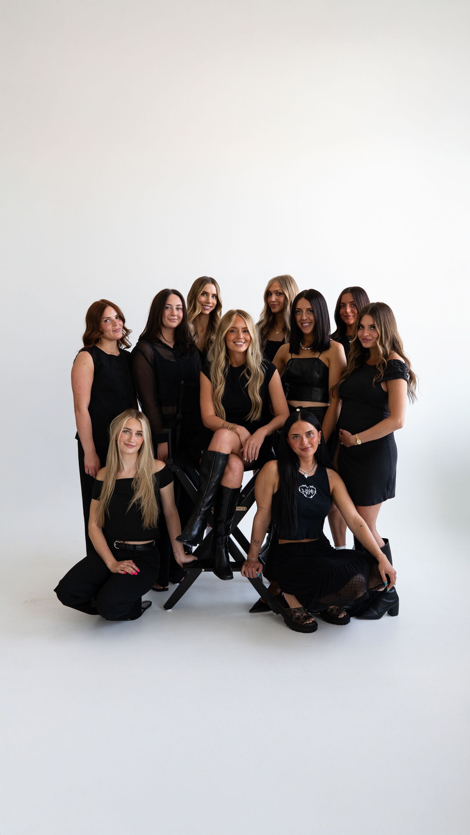 Group of ten women in coordinated black outfits posing together in a bright white photography studio.