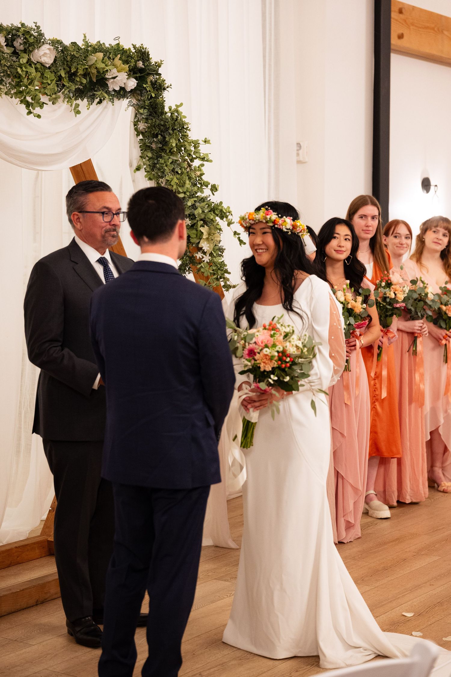 Bride in white gown and floral crown smiles at groom during indoor wedding ceremony with bridesmaids in coral dresses.