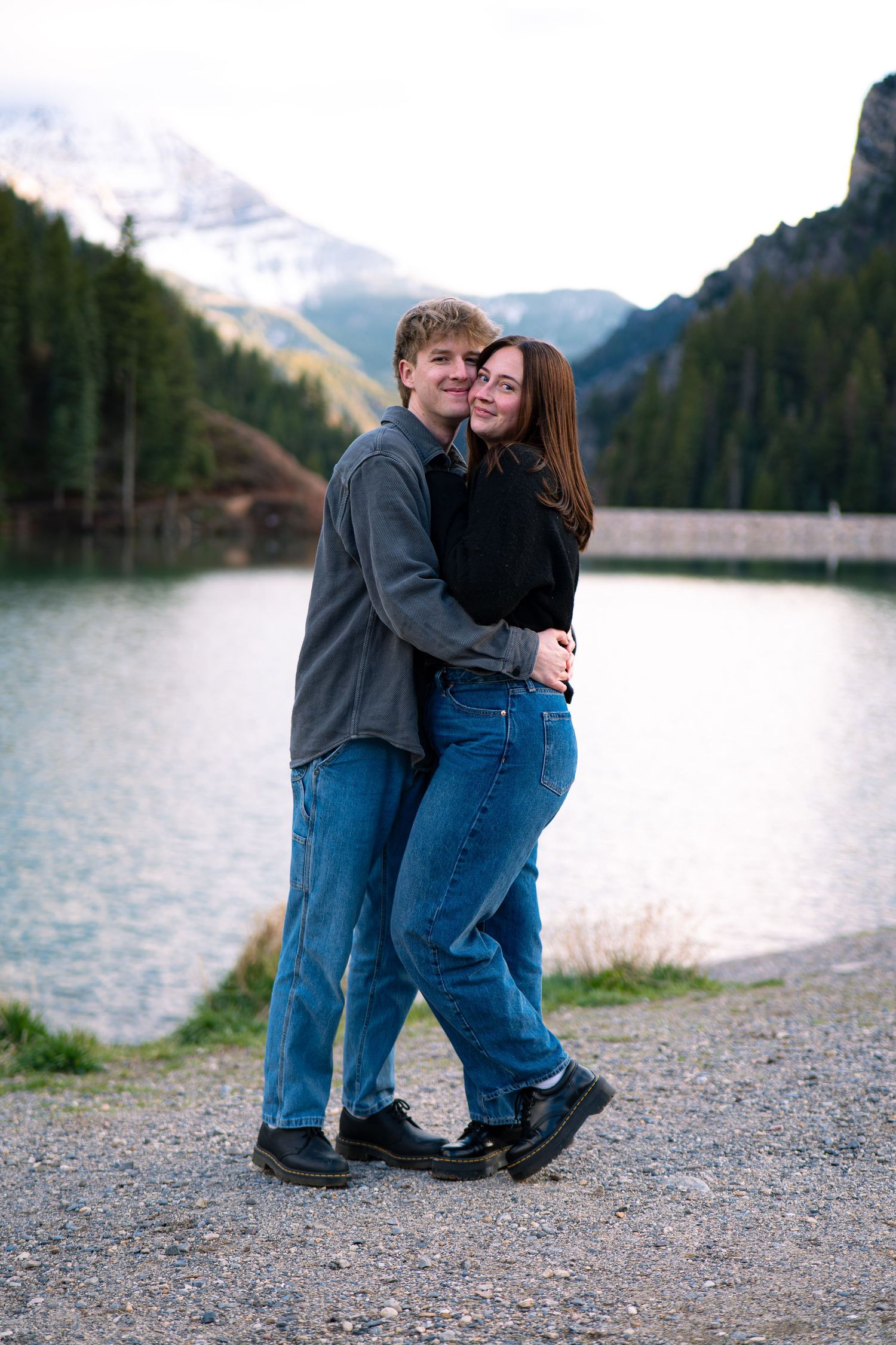 Couple embracing and smiling by a mountain lake with snow-capped peaks and pine trees in the background.
