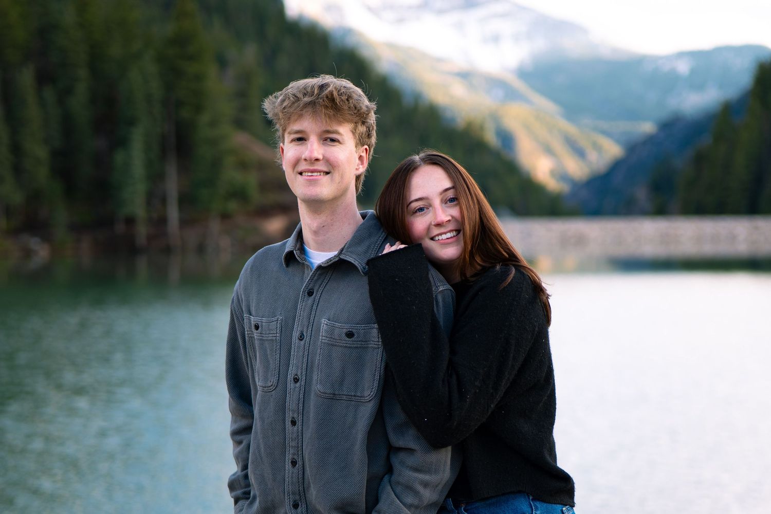 Two smiling teenagers pose together near a mountain lake with pine trees and snow-capped peaks in the background.