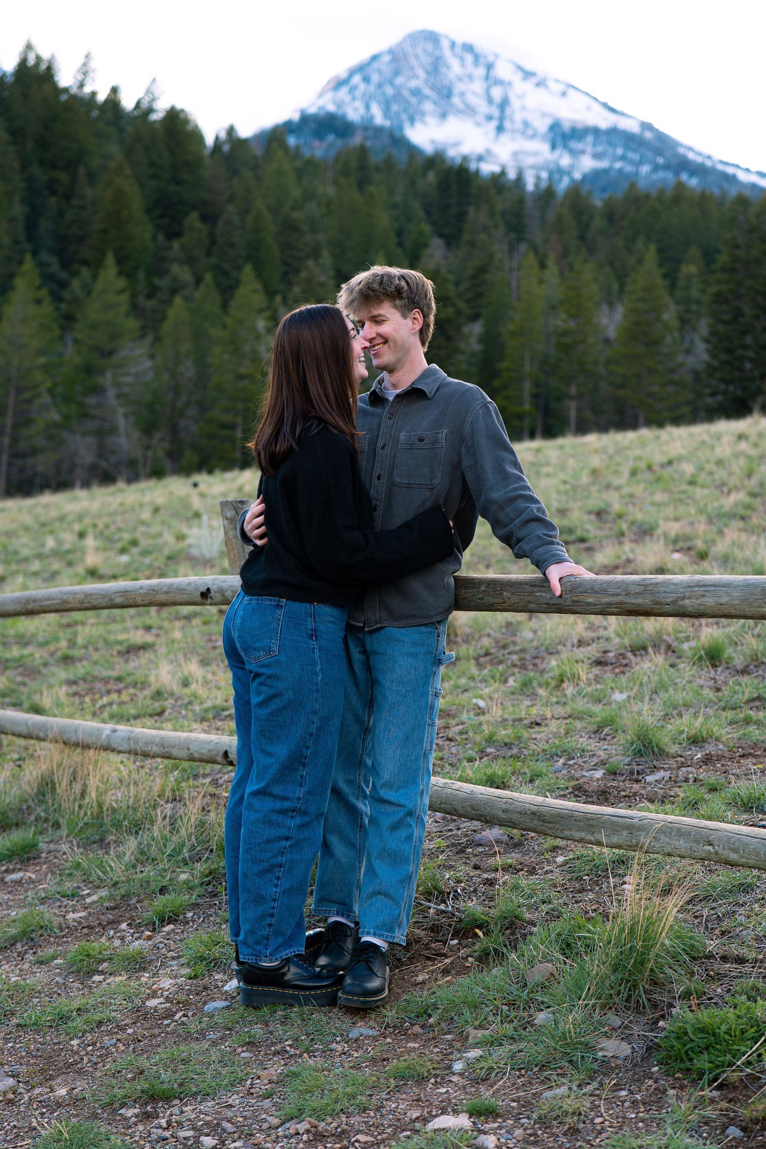 Couple embracing by a rustic wooden fence with snow-capped mountain and pine forest backdrop.