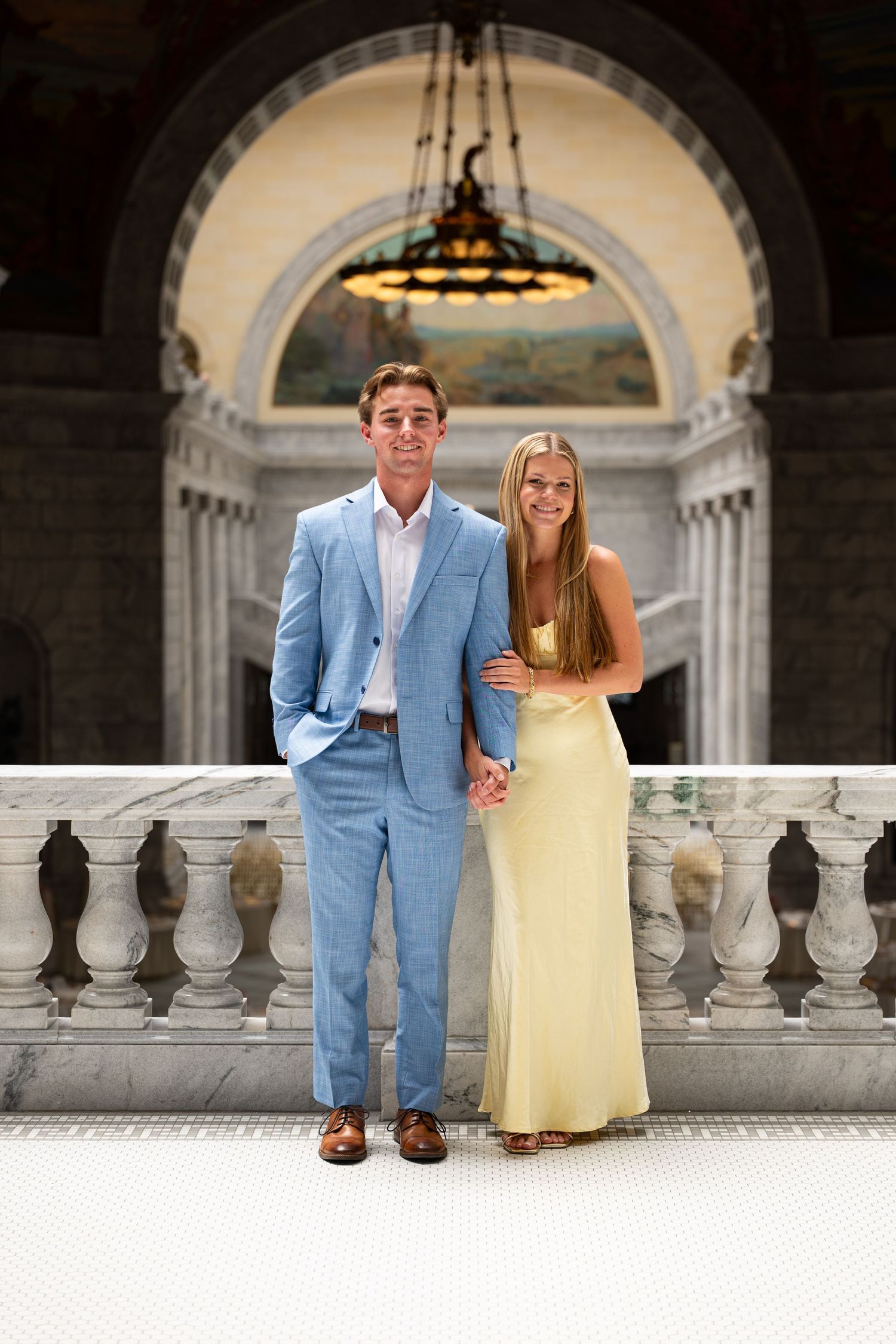 Couple posing on marble balcony in grand building, man in blue suit and woman in yellow dress.