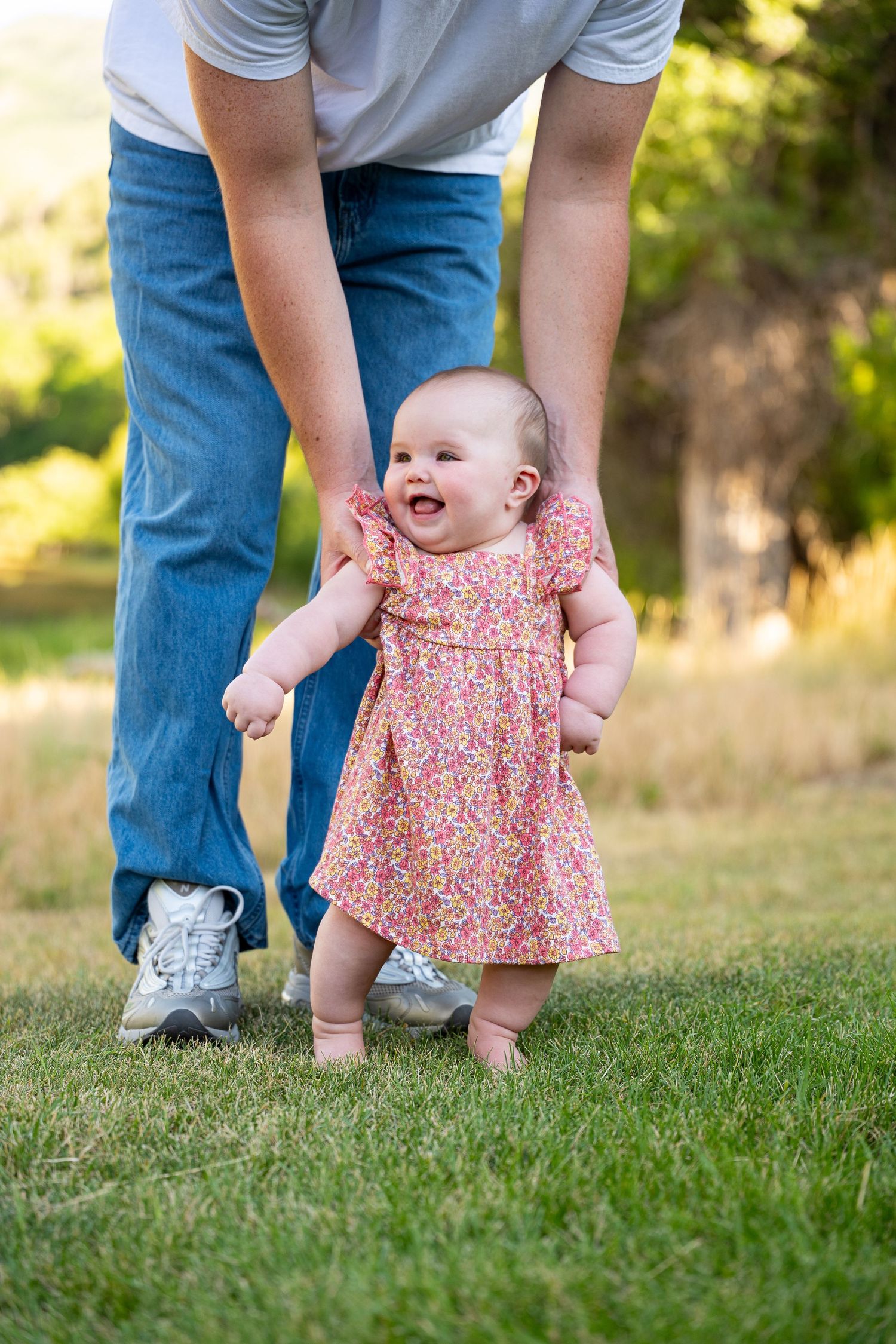 Smiling baby girl in floral dress takes first steps on grass, supported by adult hands outdoors.