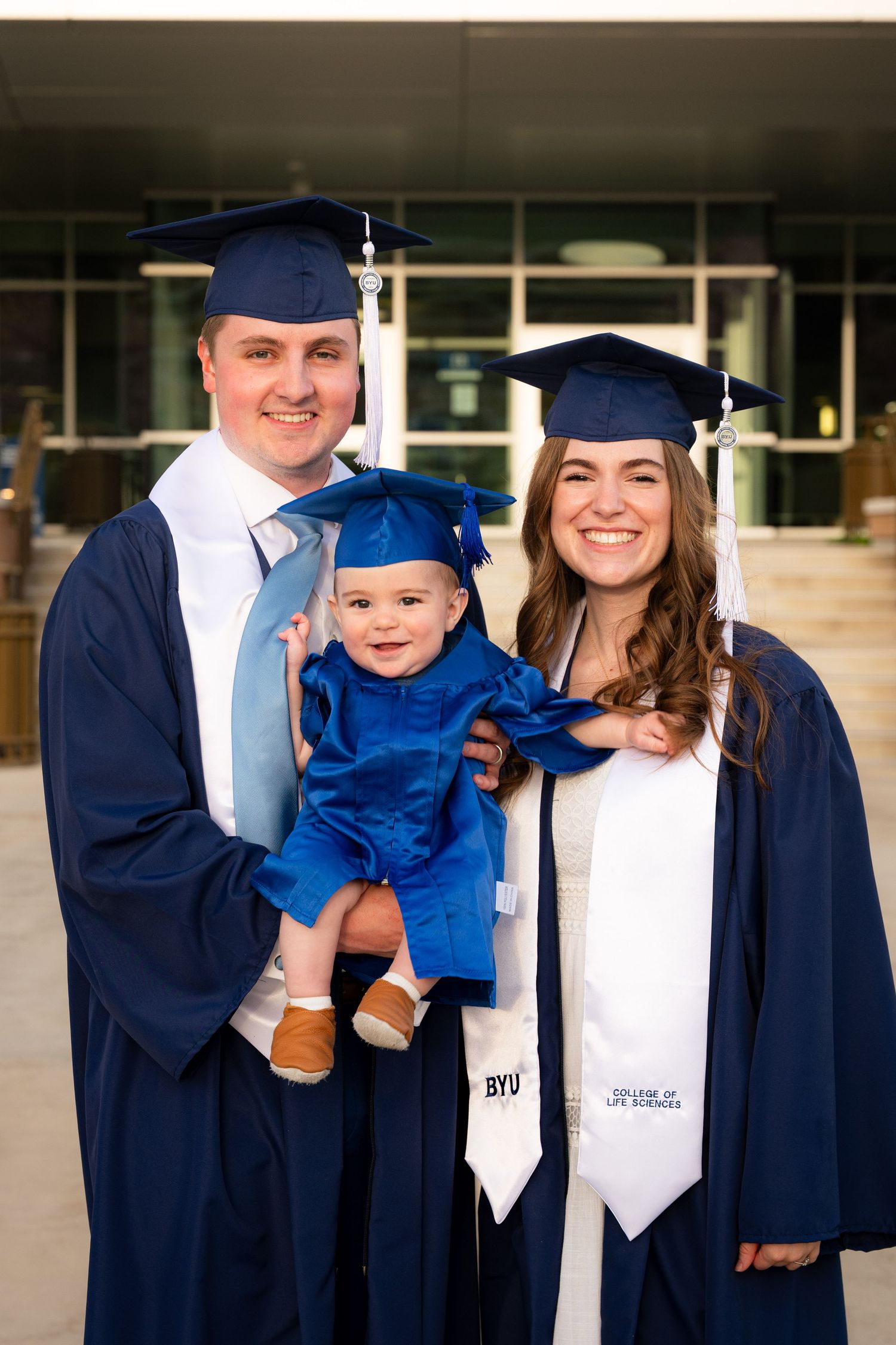A couple in graduation gowns and caps stand together in front of a university building.
