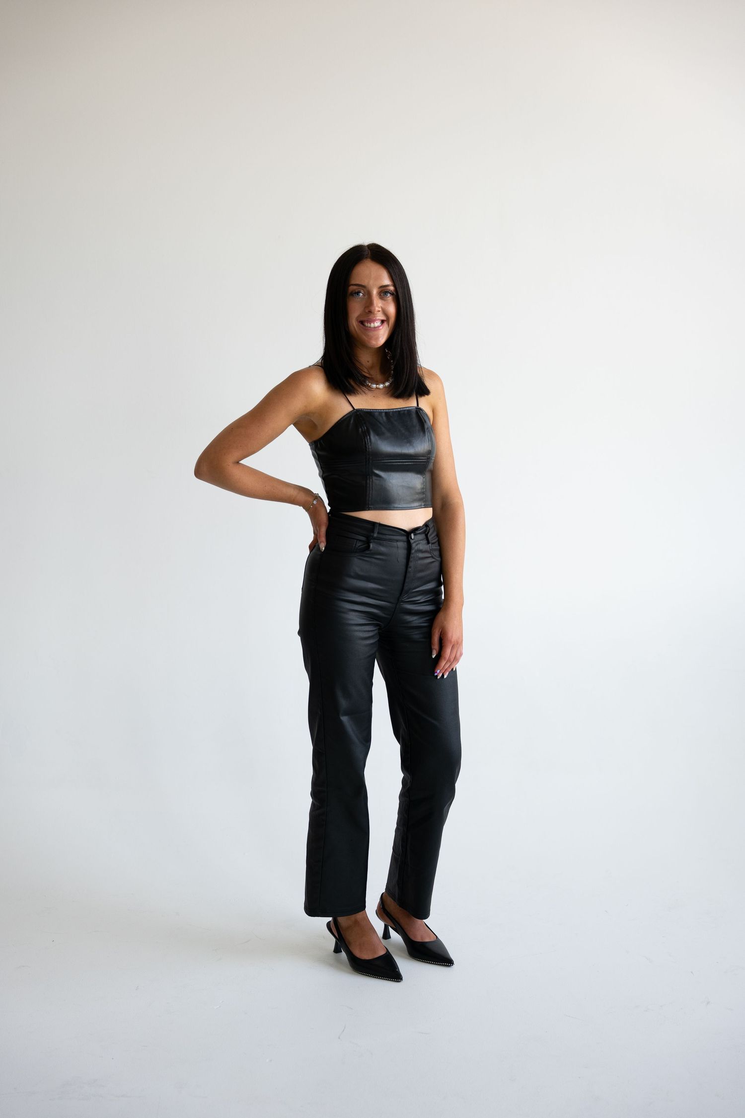 Woman in black leather crop top and coated straight-leg pants posing confidently in a white studio.