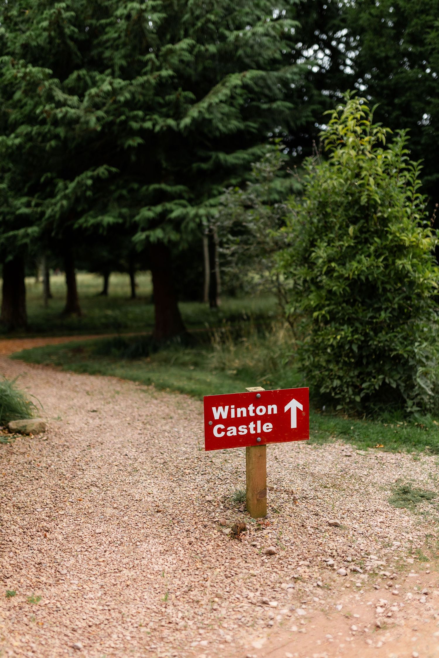 A series of trail markers with red arrows pointing to Winton Castle along a gravel path through wooded grounds.