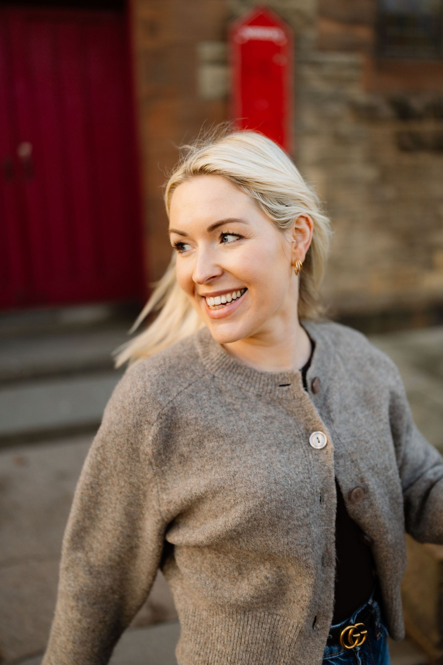 Person in grey knit sweater with designer accessory smiling against brick wall.
