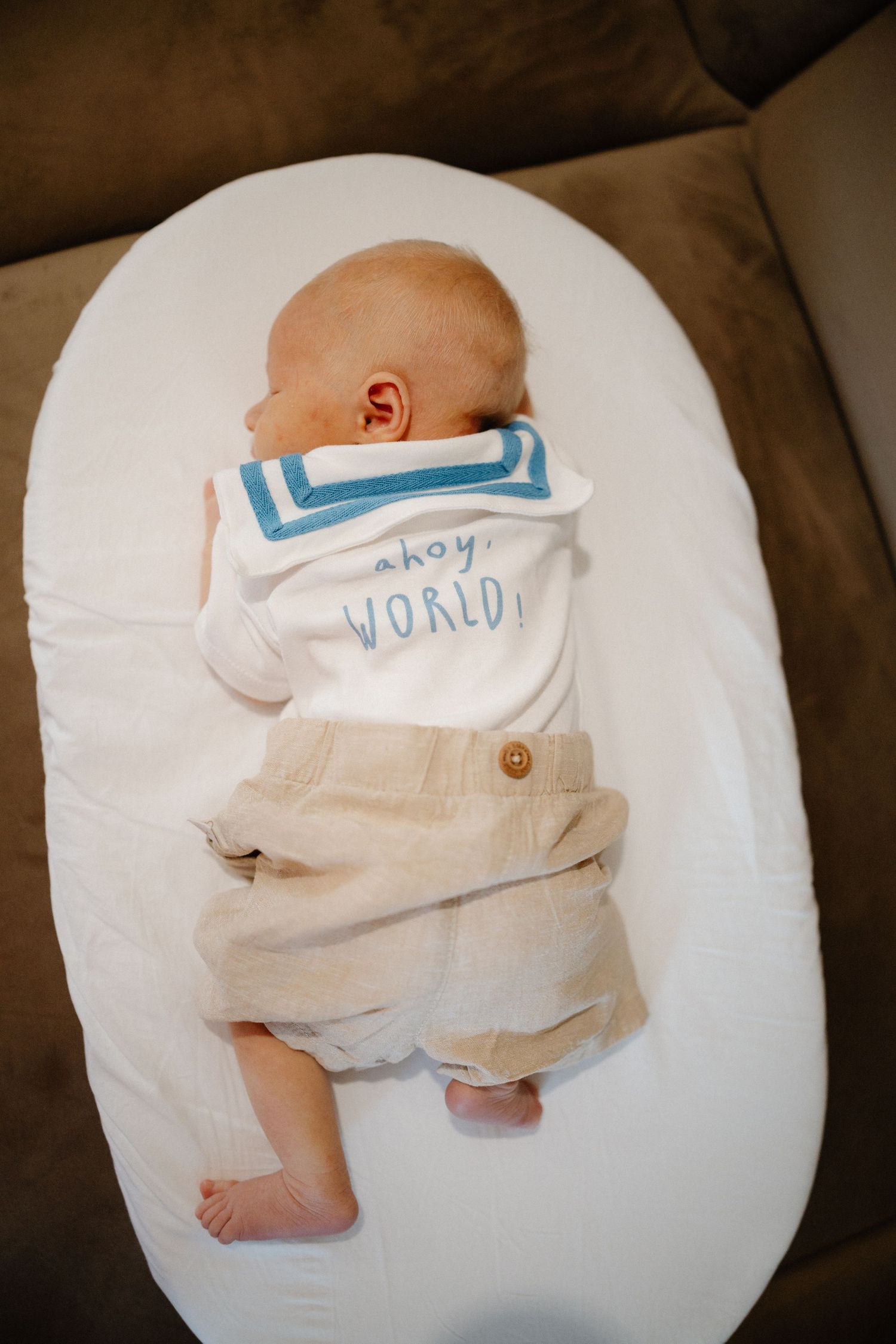 Baby wearing sailor-style outfit lying on white cushion, viewed from above.