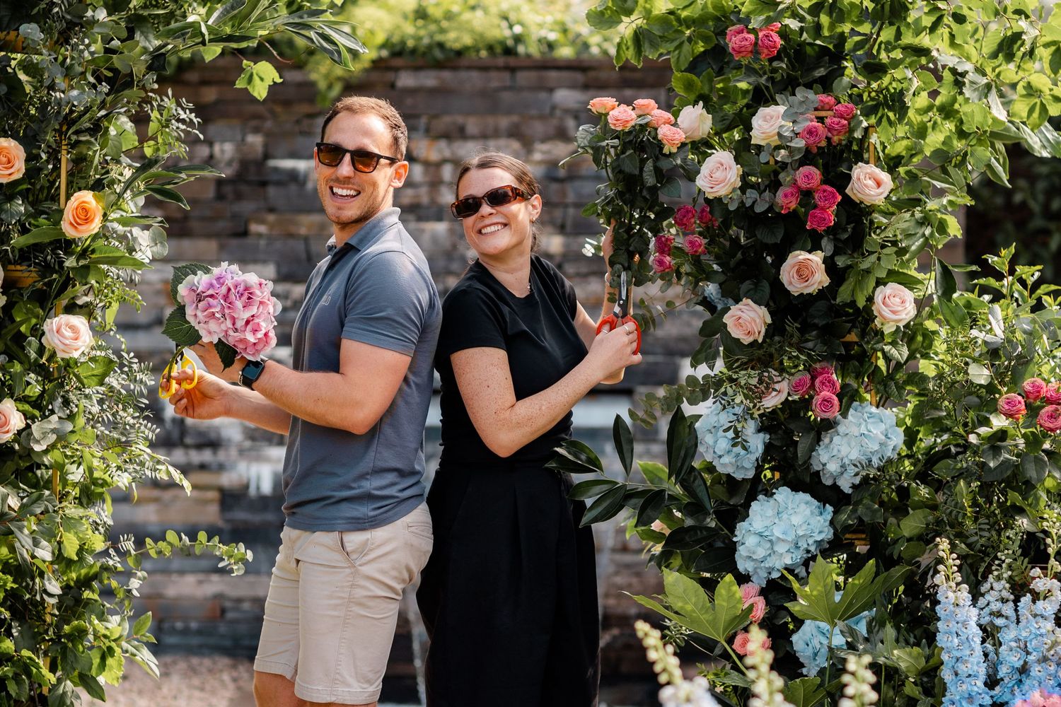 Gardeners working together among blooming flowers and hydrangeas in a lush garden setting.