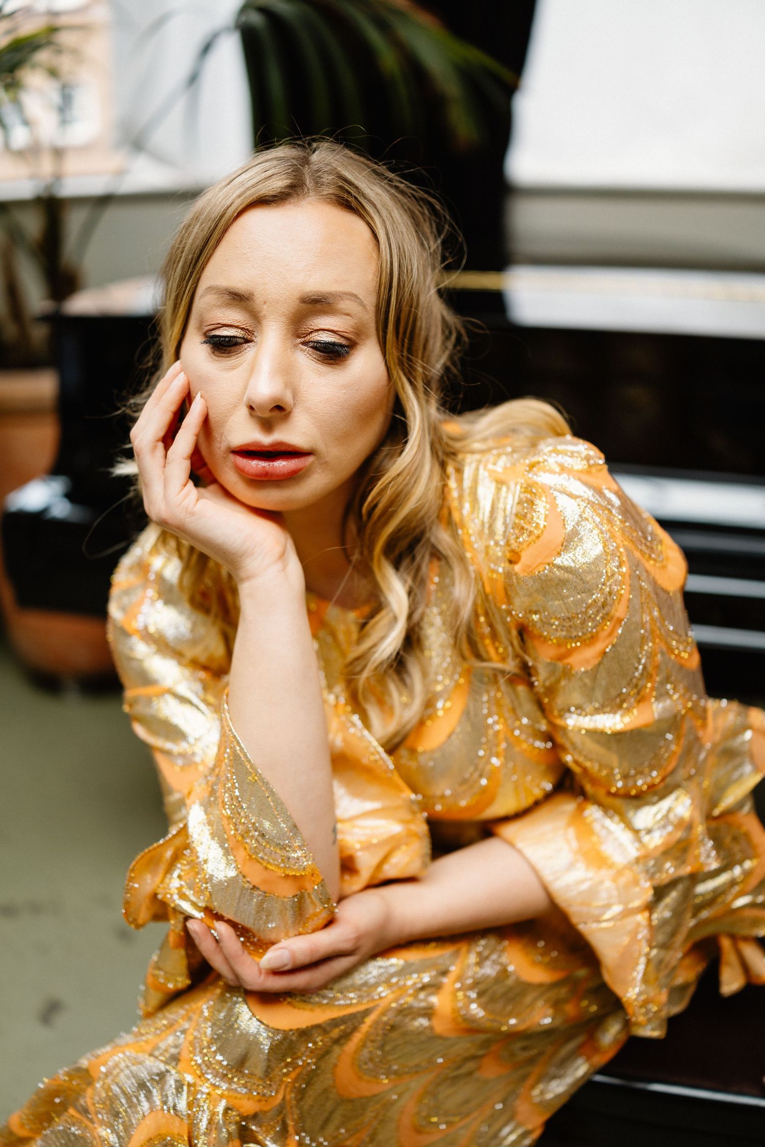 Portrait of person in gold sequined dress resting head on hand near piano and plants.