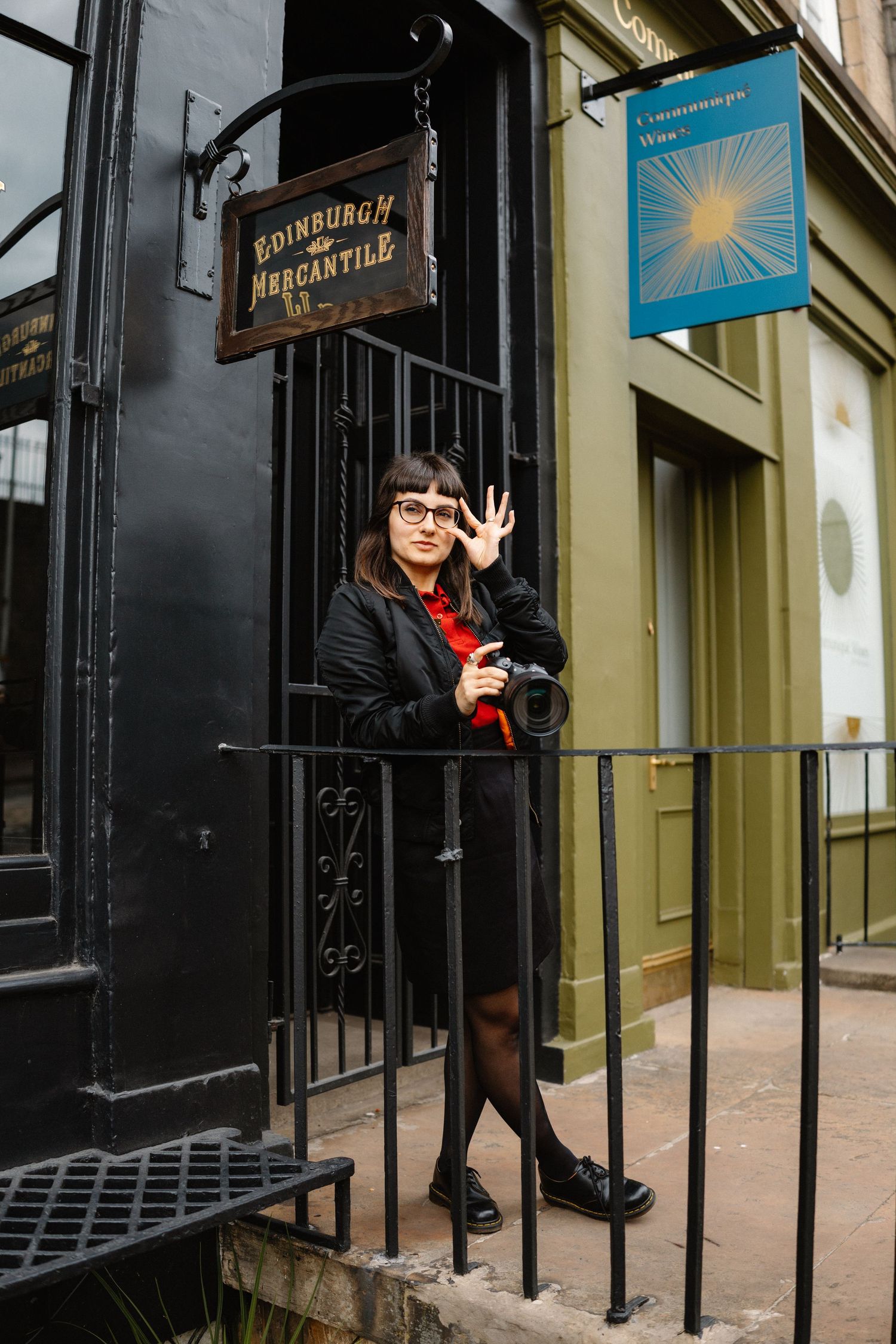 Photographer in black clothing takes pictures near a storefront with Edinburgh signage.