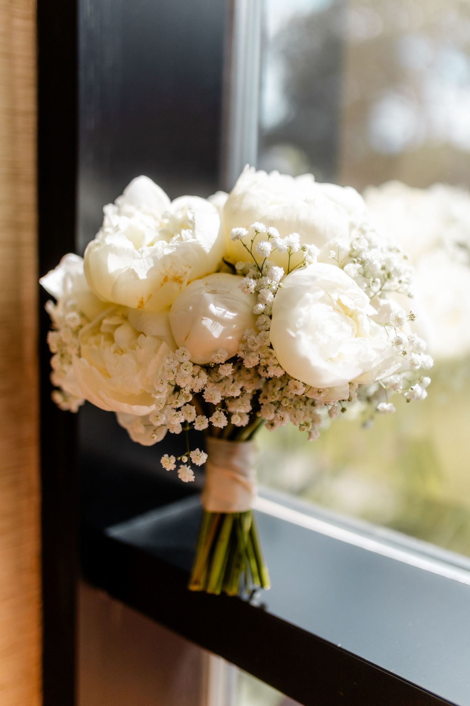 A delicate white wedding bouquet with peonies and baby's breath sits on a windowsill in soft natural light.