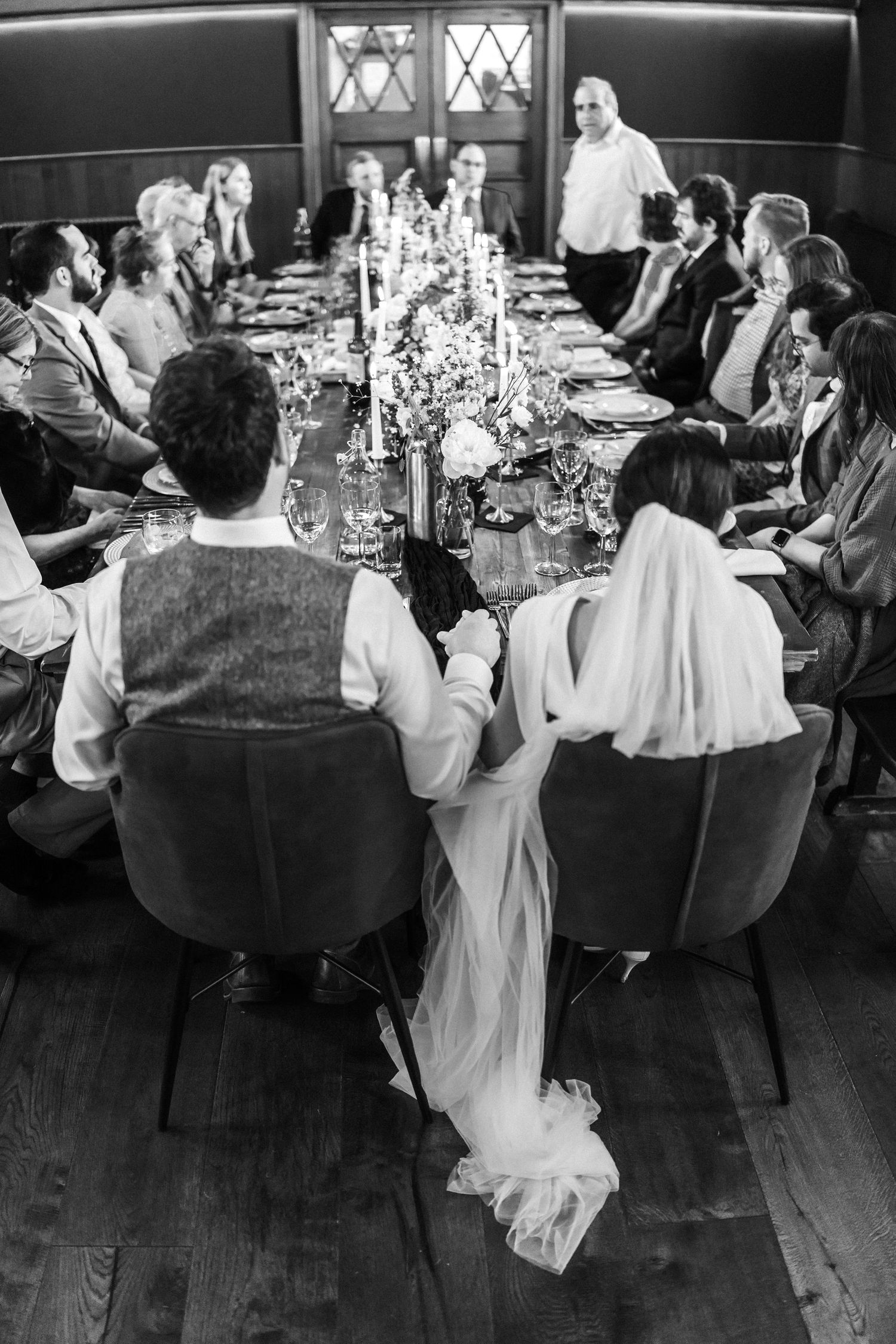 Wedding reception guests seated at elegantly decorated tables with candlelight in a dark wood-paneled room.