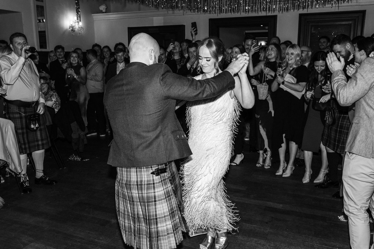 A couple dancing at a reception with a crowd of guests in the background.