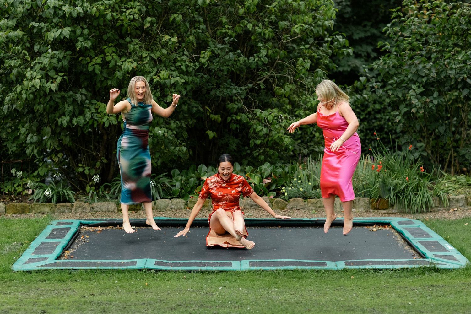 Three people in formal attire jumping on a large outdoor trampoline surrounded by greenery.