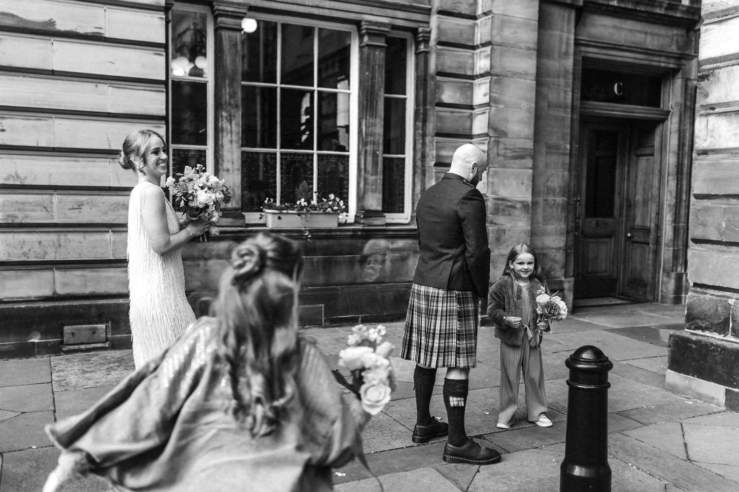 Wedding couple embraces outside historic stone building with traditional Scottish attire.