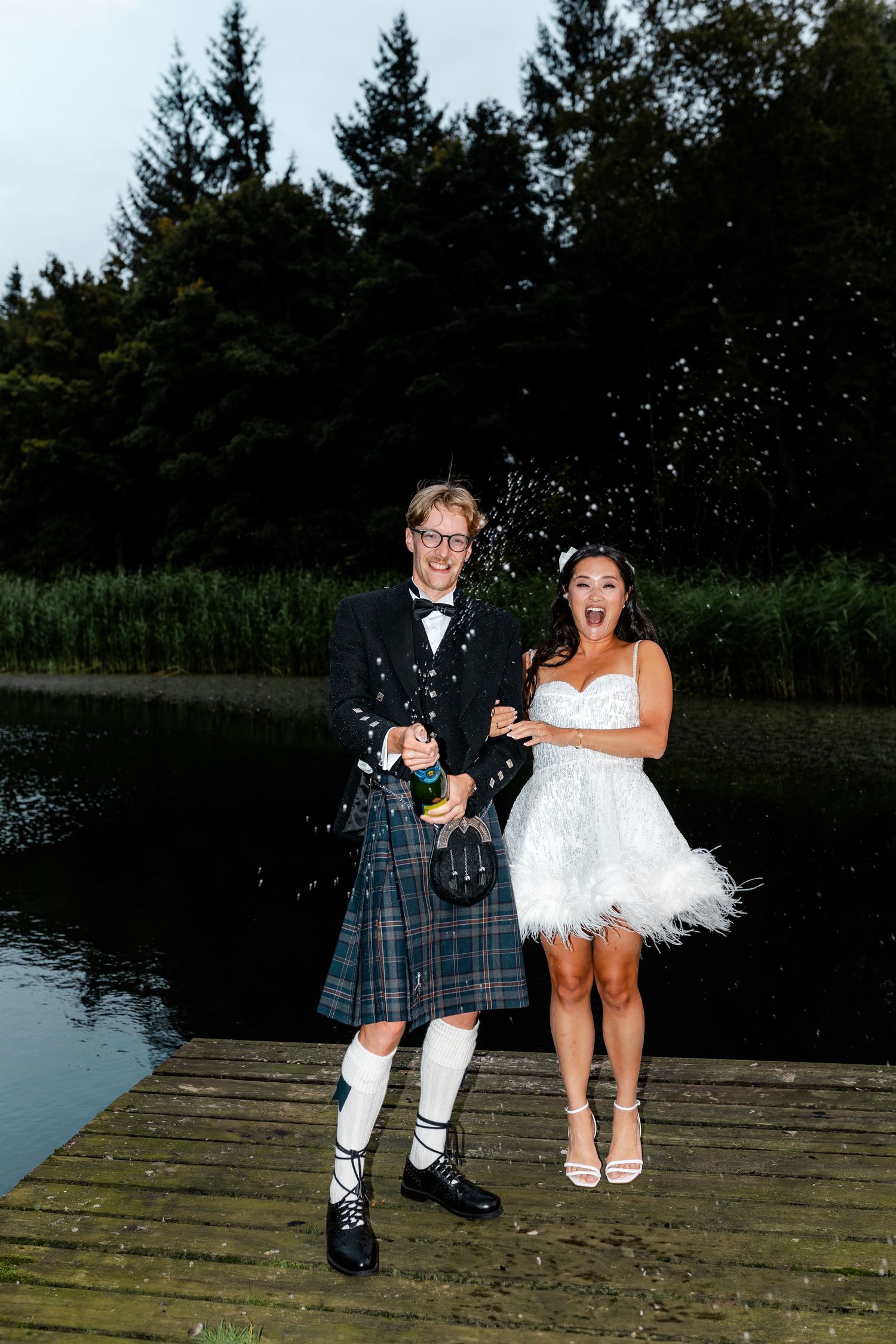 Couple in Scottish wedding attire posing on wooden dock by lake.