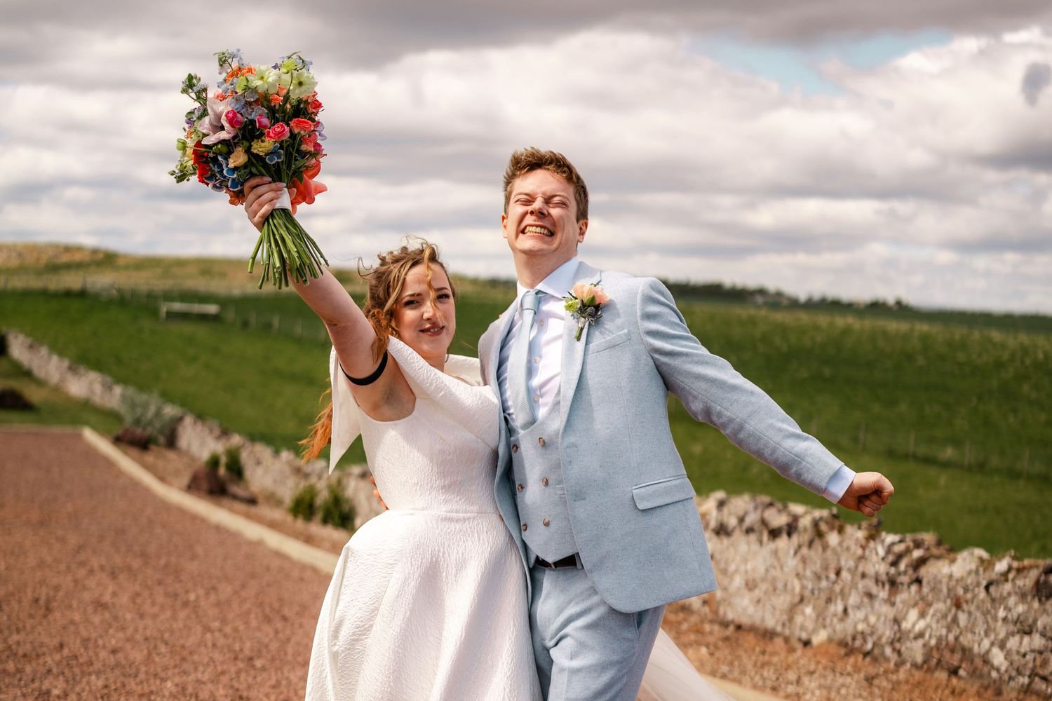 Couple celebrates joyfully with raised bouquet against cloudy countryside backdrop.