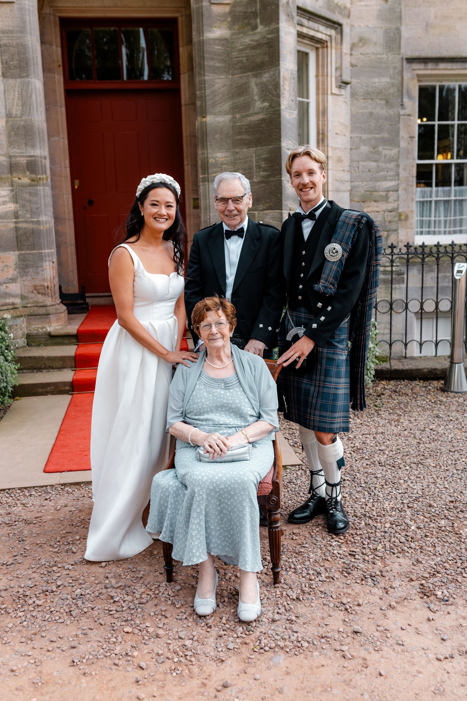 Bride in white gown and groom in Scottish kilt pose with family outside a stone building.