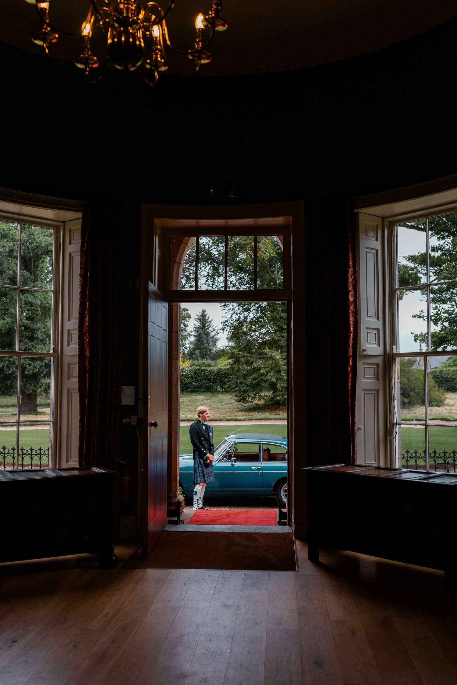 A groom stands in an elegant doorway of a grand estate, with a vintage teal car visible on the red carpet outside.