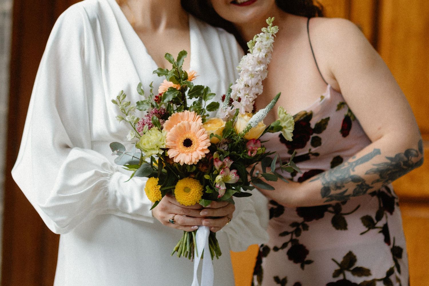 Two women holding a colorful bridal bouquet with peach gerberas, yellow flowers, and greenery at a wedding.
