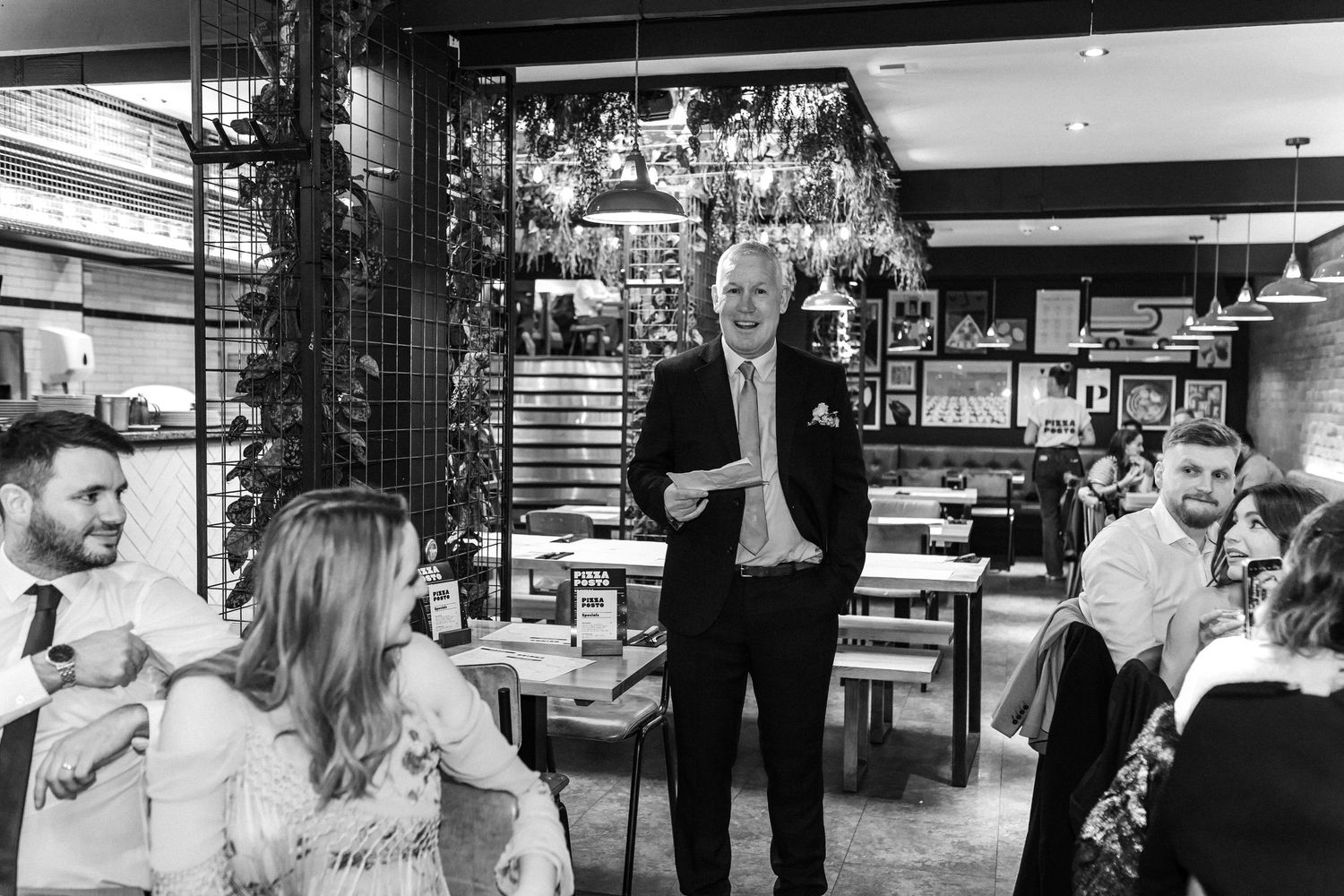 Elderly man giving a speech to wedding guests in a modern restaurant in black and white photo.