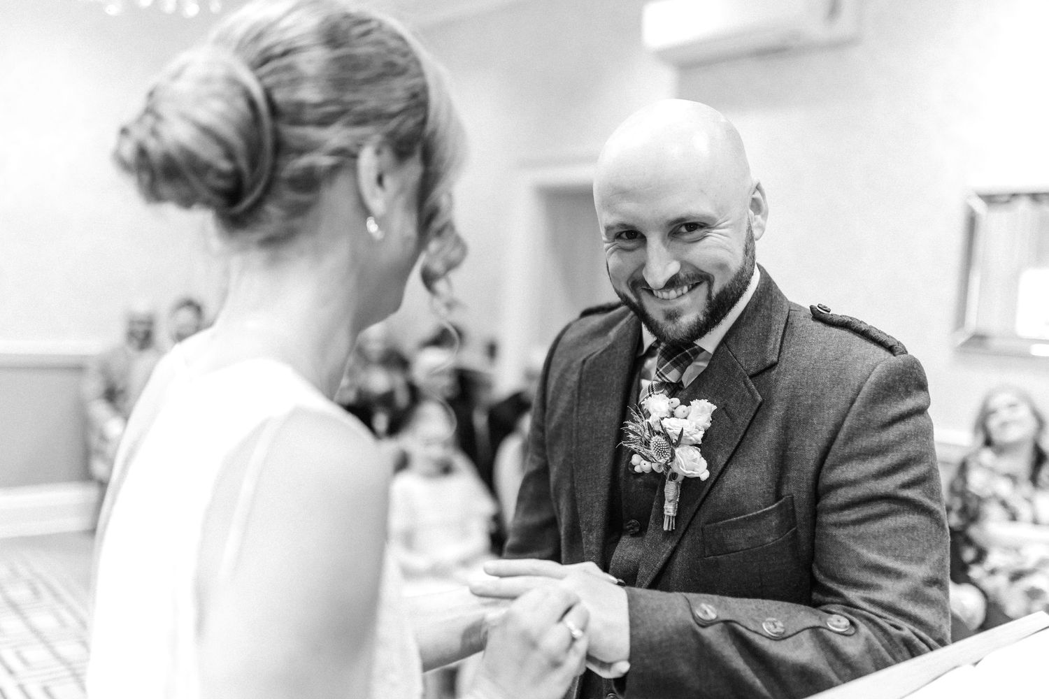 Groom in tartan kilt smiling warmly at bride during wedding ring exchange ceremony in black and white.