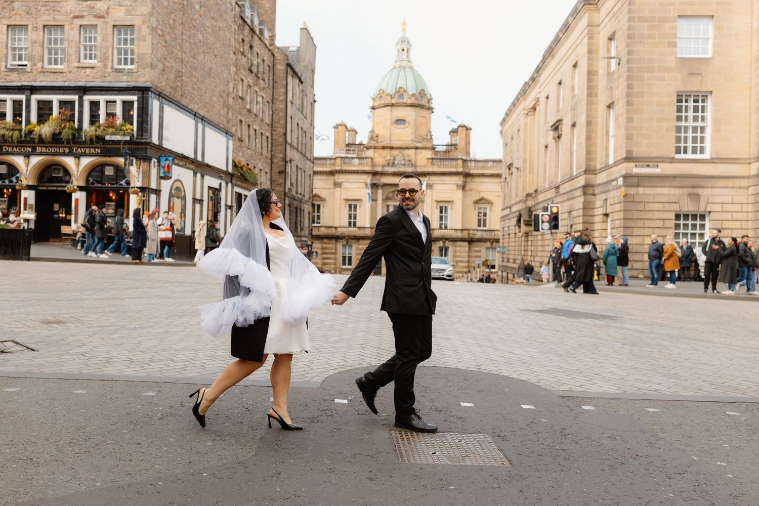 Bride and groom holding hands crossing a cobblestone square in Edinburgh, bride wearing short dress with tulle veil.