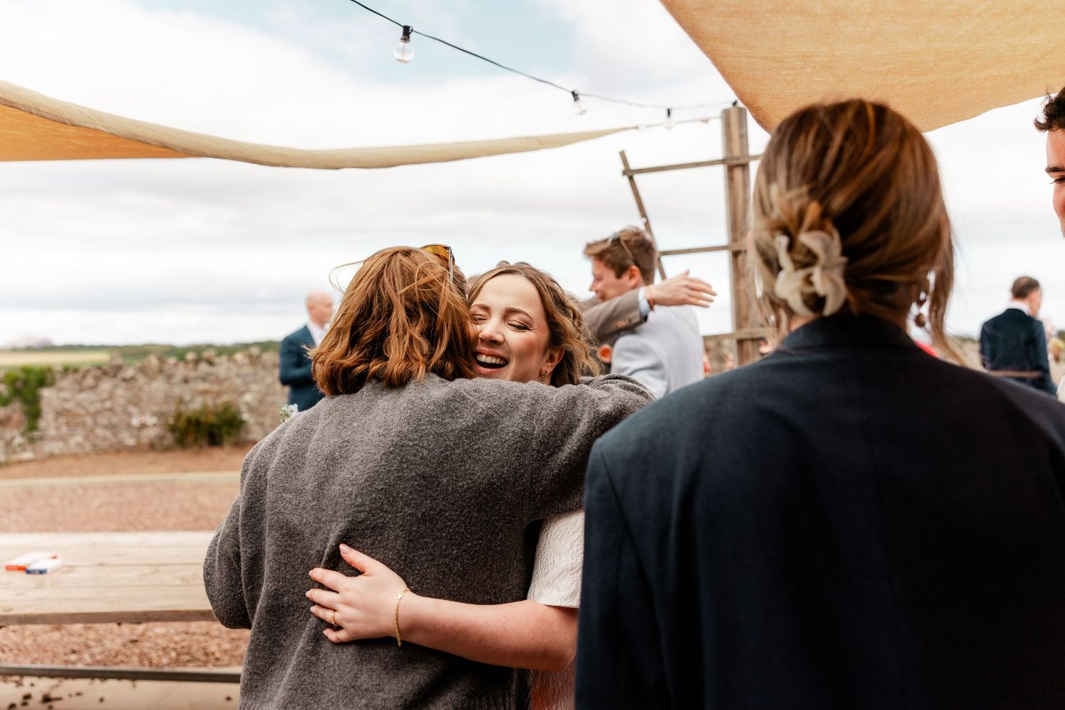 People hugging and laughing at an outdoor gathering under a canopy with string lights.