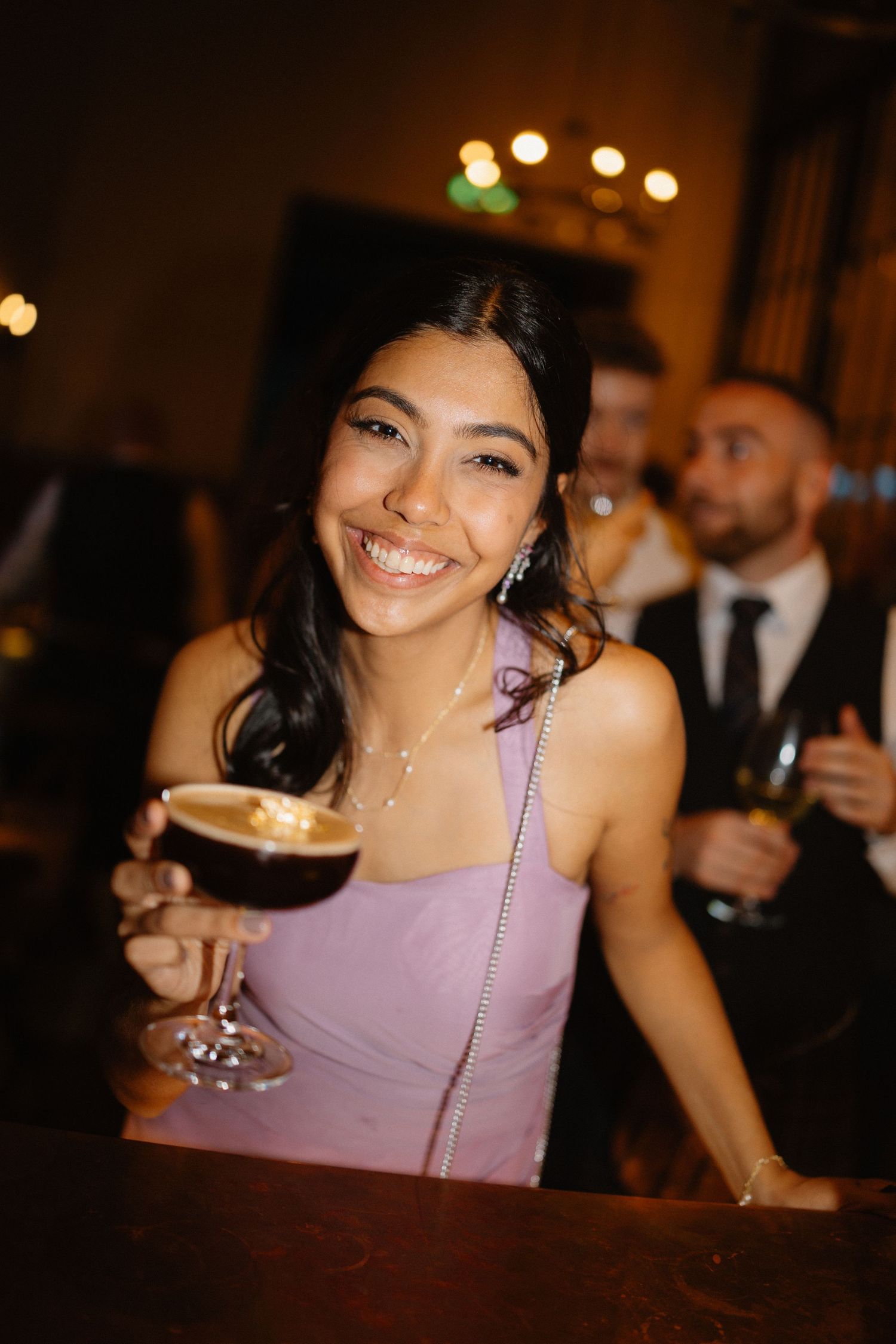 Smiling wedding guest in lavender dress holds espresso martini at warmly lit reception bar.