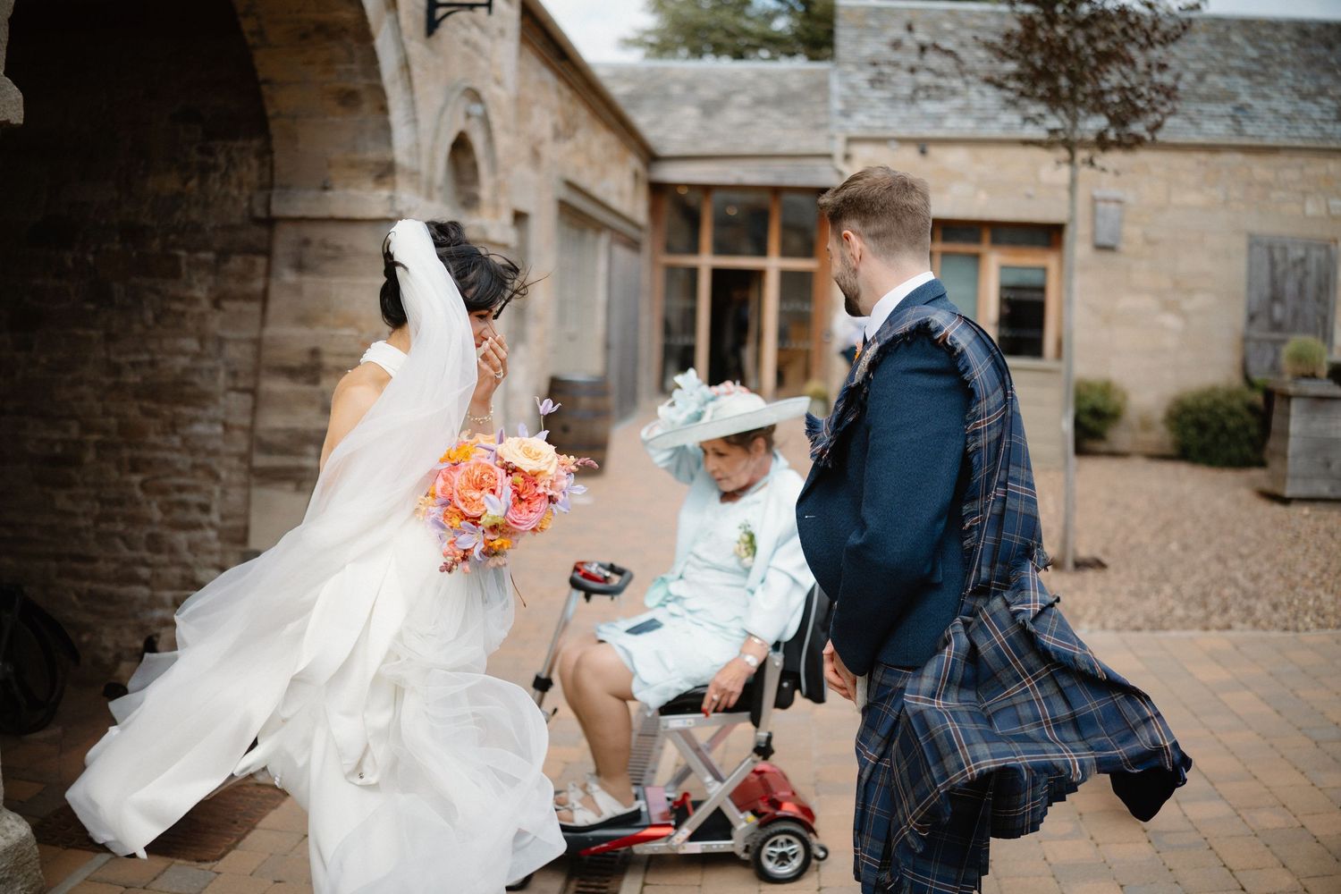 Bride in white gown with bouquet greets elderly guest in mobility scooter outside stone wedding venue.