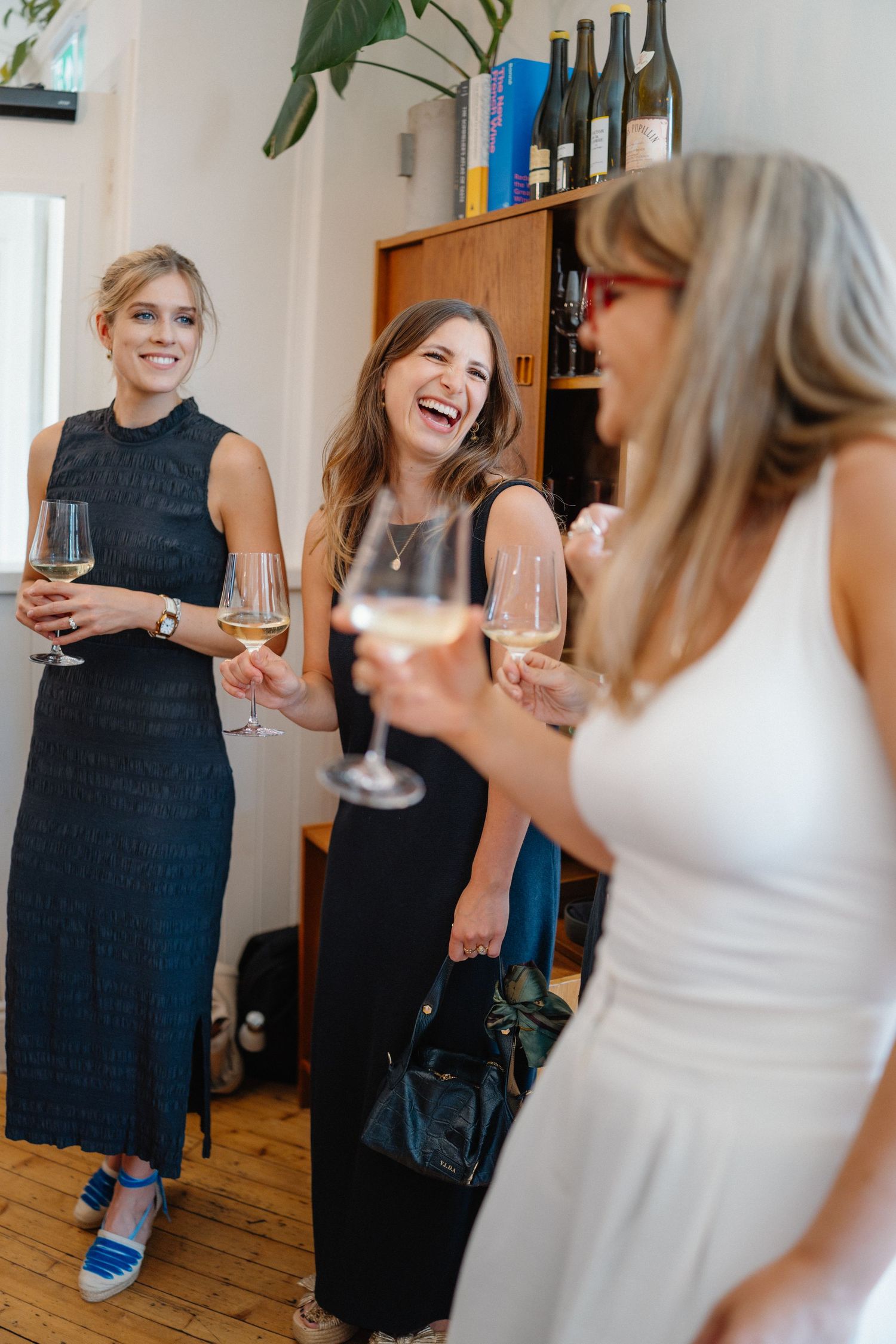 Three women in elegant dresses laughing and holding wine glasses at a stylish indoor gathering.