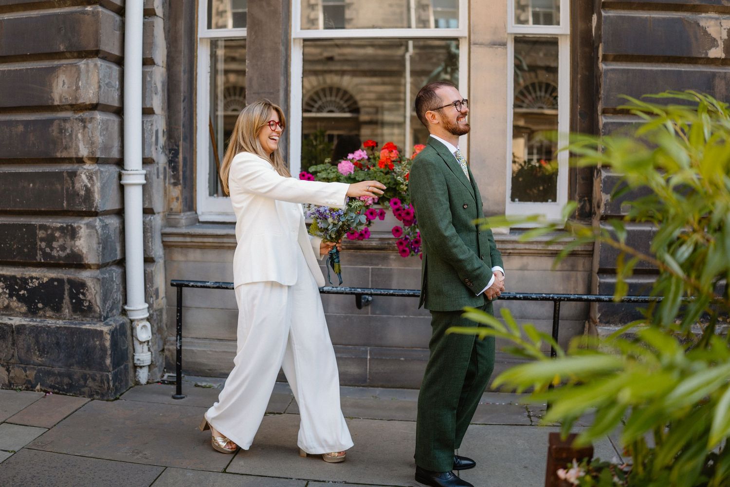 Bride in white suit hands flowers to groom in green suit outside a stone building on their wedding day.