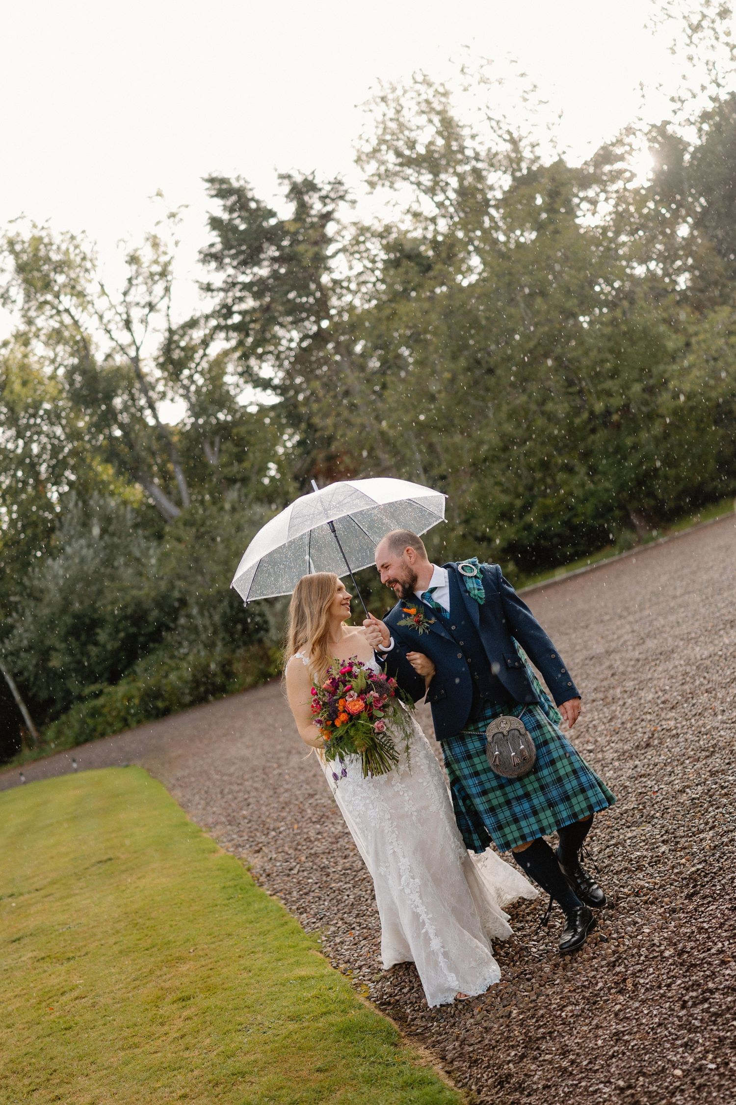 Bride and groom share an intimate moment under a clear umbrella in the rain holding a colorful bouquet.