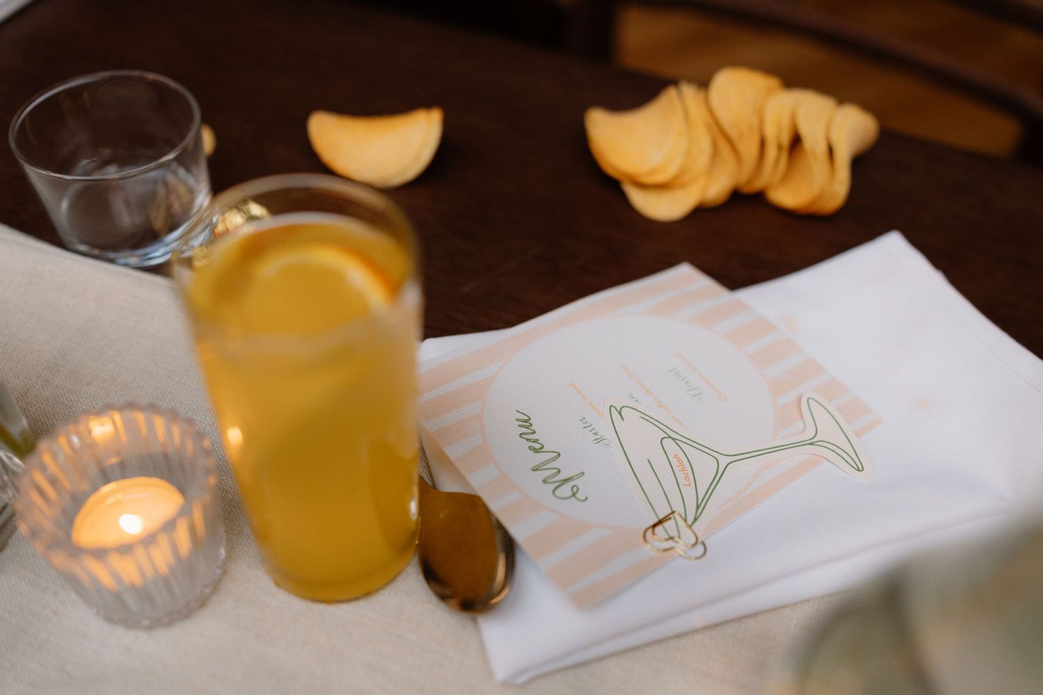 Wedding menu card beside a glass of orange juice, candle, and snacks on a linen-covered reception table.
