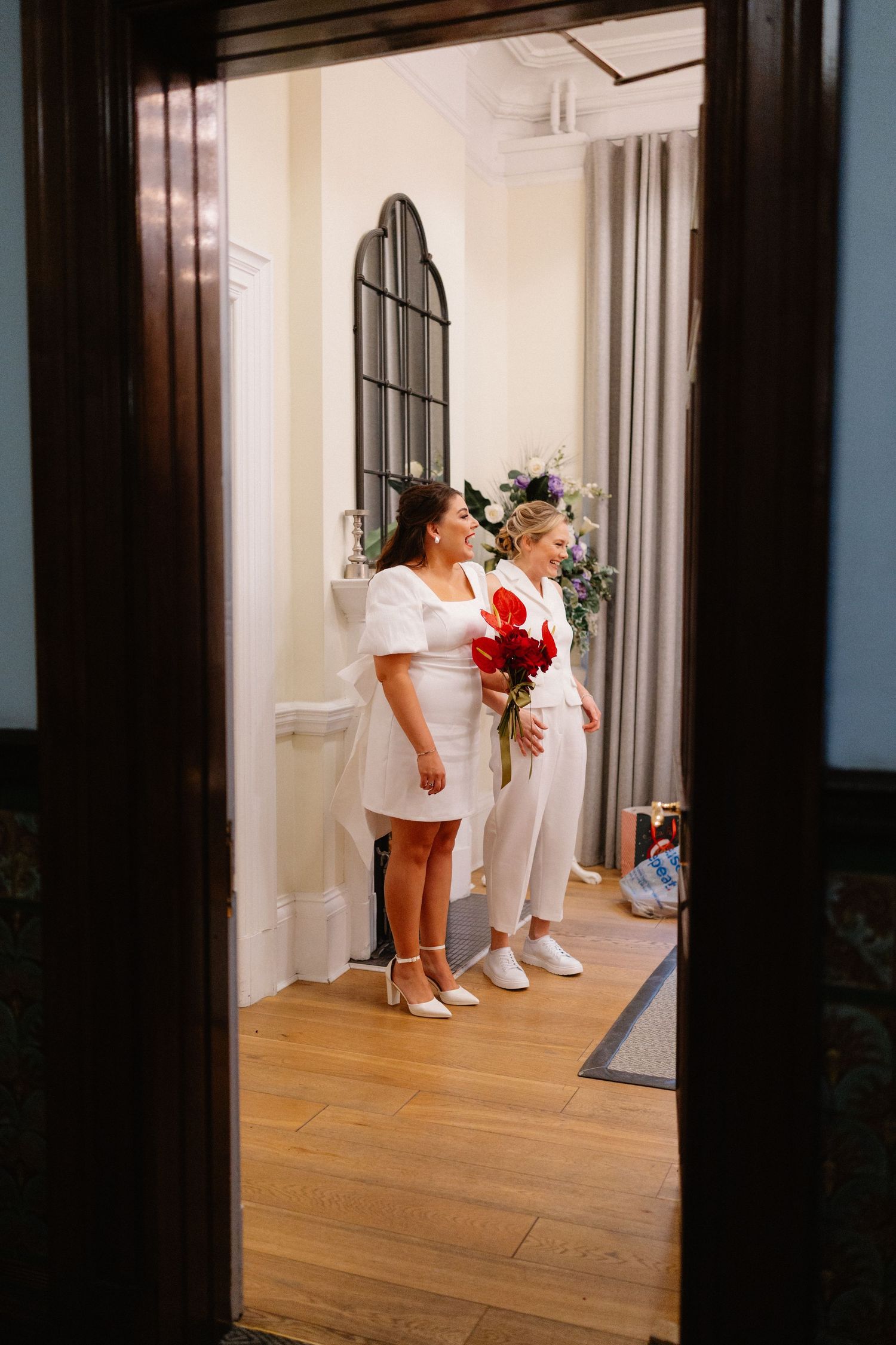 Two brides in white outfits holding red floral bouquets stand in a elegant room viewed through a doorway.