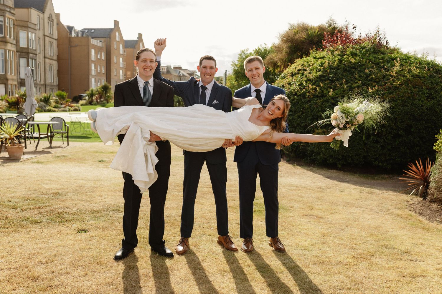 Groomsmen in navy suits lift smiling bride in white wedding dress outdoors on green lawn beside elegant building.