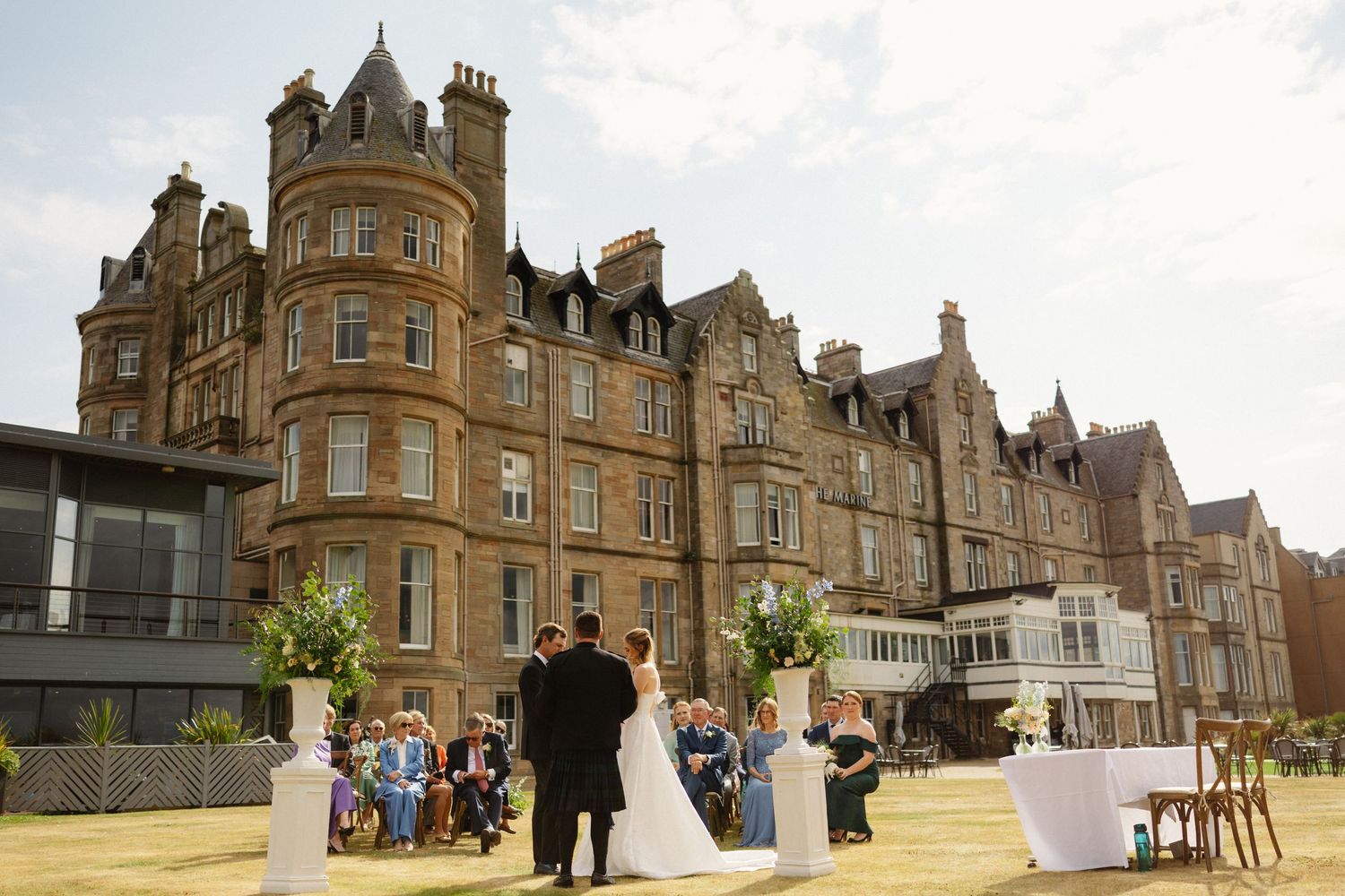 Elegant outdoor wedding ceremony on manicured lawn in front of grand Victorian stone hotel building on sunny day.