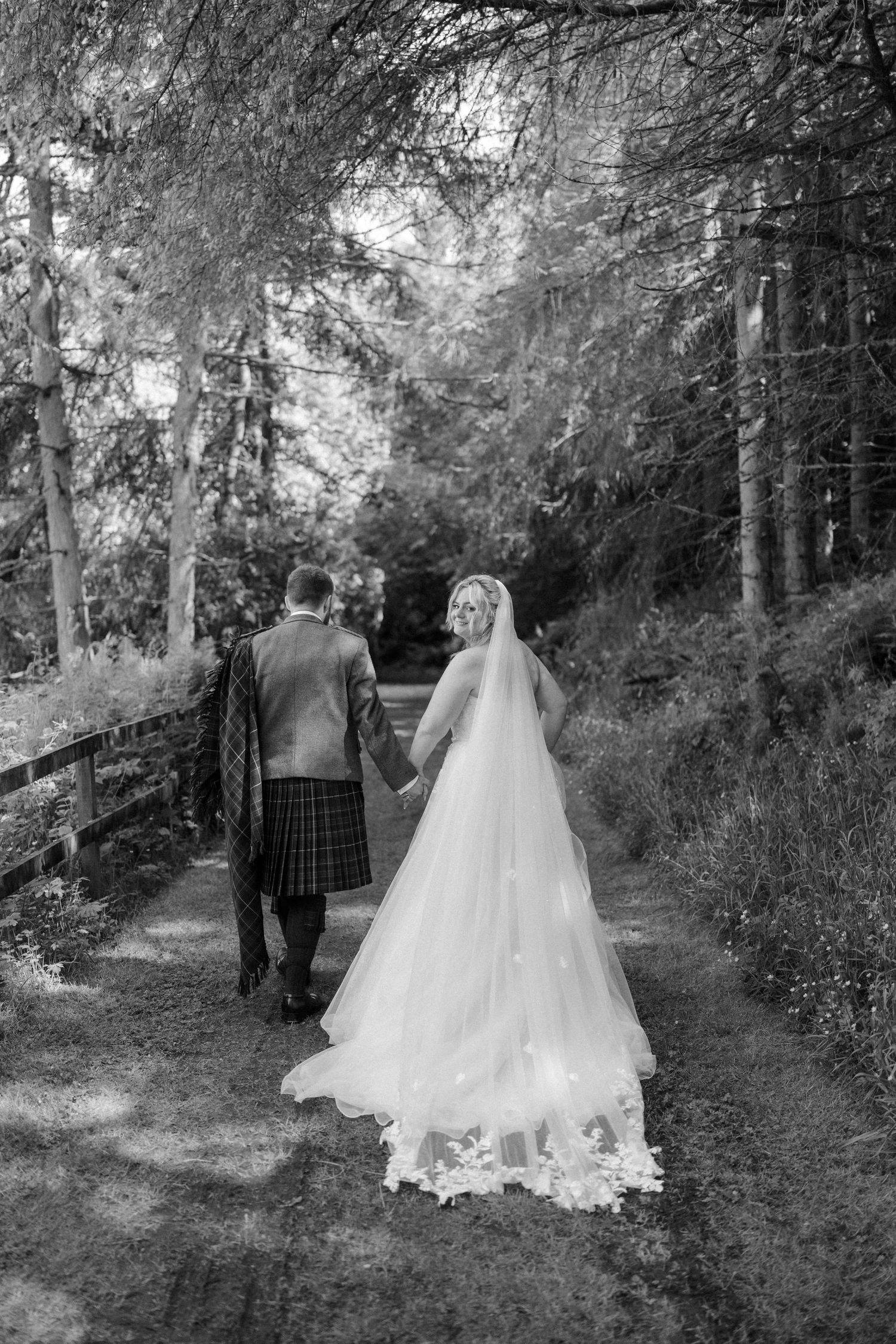 Bride in flowing gown and groom in tartan kilt walk hand-in-hand along a woodland path.