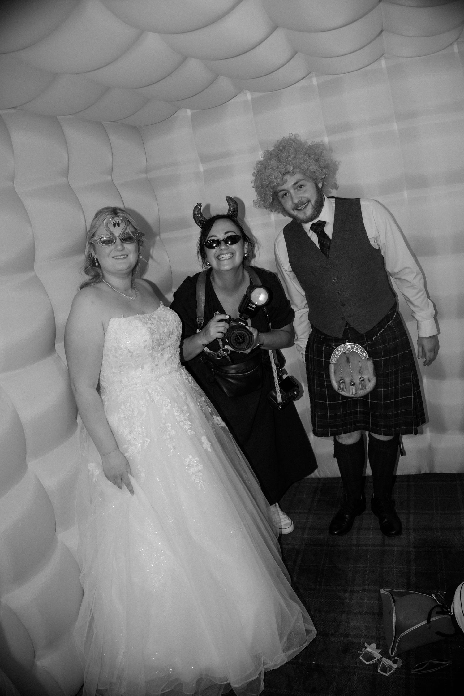 Bride and groom in Scottish kilt pose with photographer wearing devil horns in black and white photo booth.