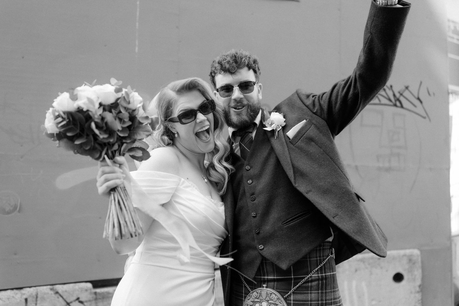 Joyful bride and groom celebrate outdoors in black and white, groom raising arm triumphantly at wedding.