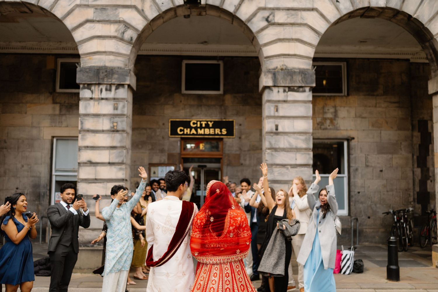 Wedding couple celebrated by guests outside Edinburgh City Chambers, bride in red lehenga, guests in colorful traditional attire.