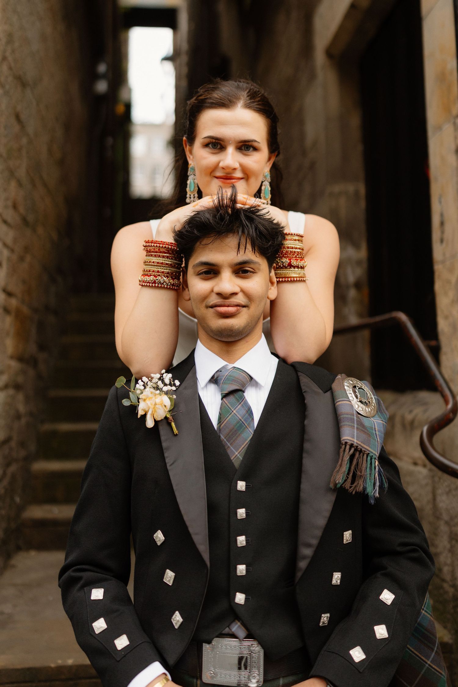 Couple in wedding attire, groom in Scottish kilt, bride wearing colorful bangles, posing in a stone alleyway.